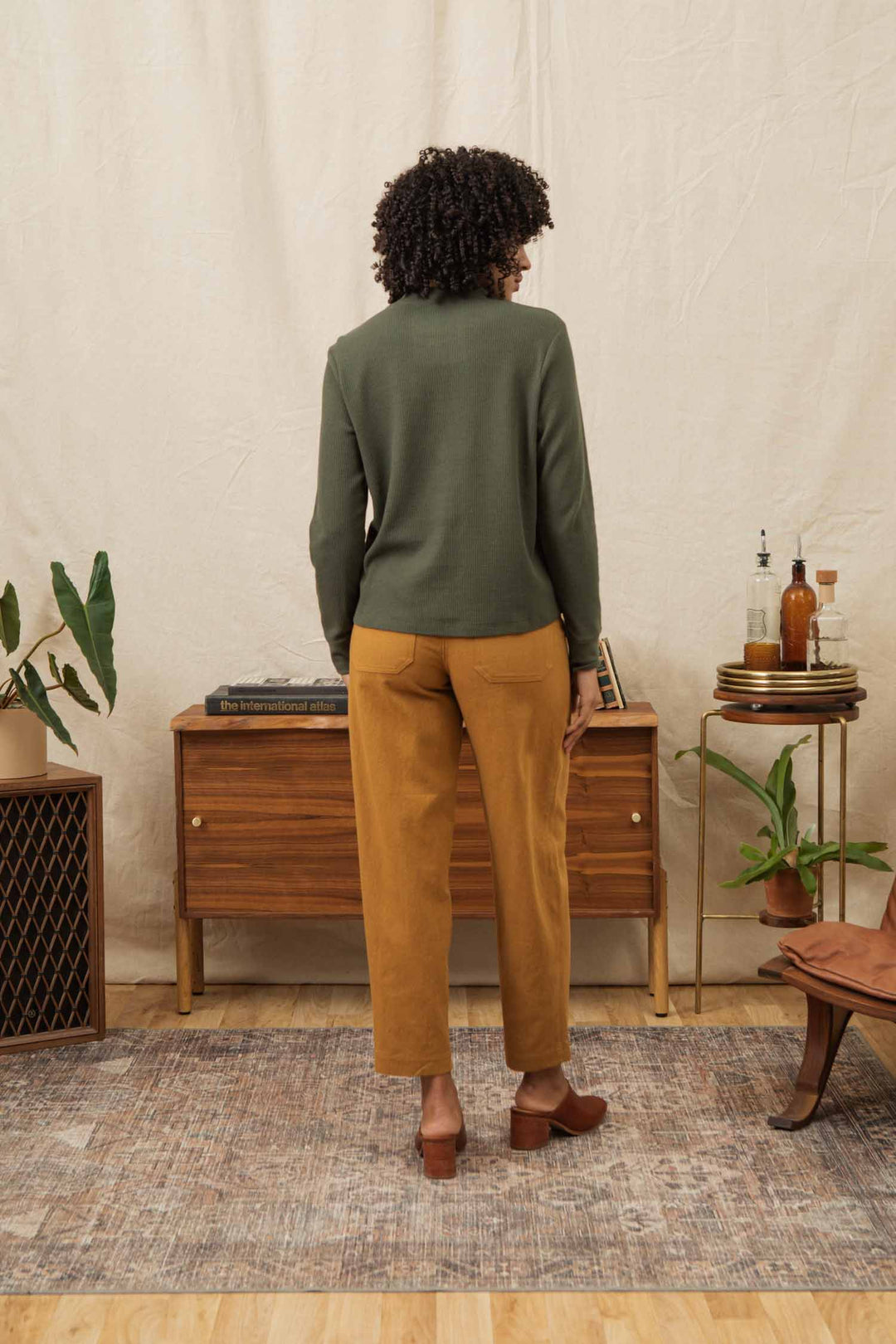 A person with curly hair, wearing the Bridge & Burn Women's Recycled Waffle Henley in Forest Melange and mustard yellow pants, stands facing away in a cozy room with wooden furniture, plants, and a patterned rug.