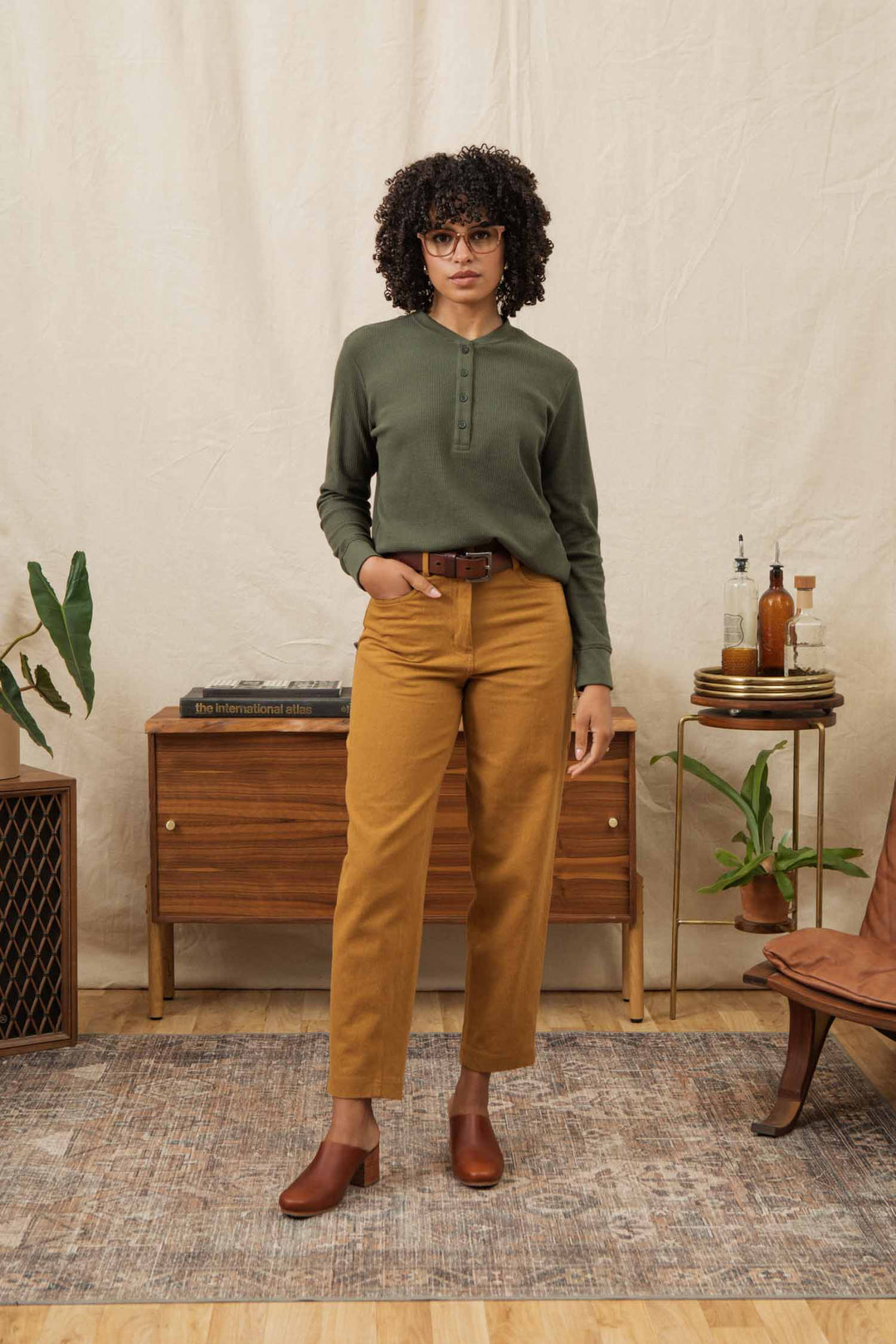 A woman with curly hair and glasses stands indoors wearing the Bridge & Burn Women's Recycled Waffle Henley in Forest Melange, paired with mustard-yellow pants and brown shoes, posing before a wooden sideboard adorned with books, plants, and glass bottles.