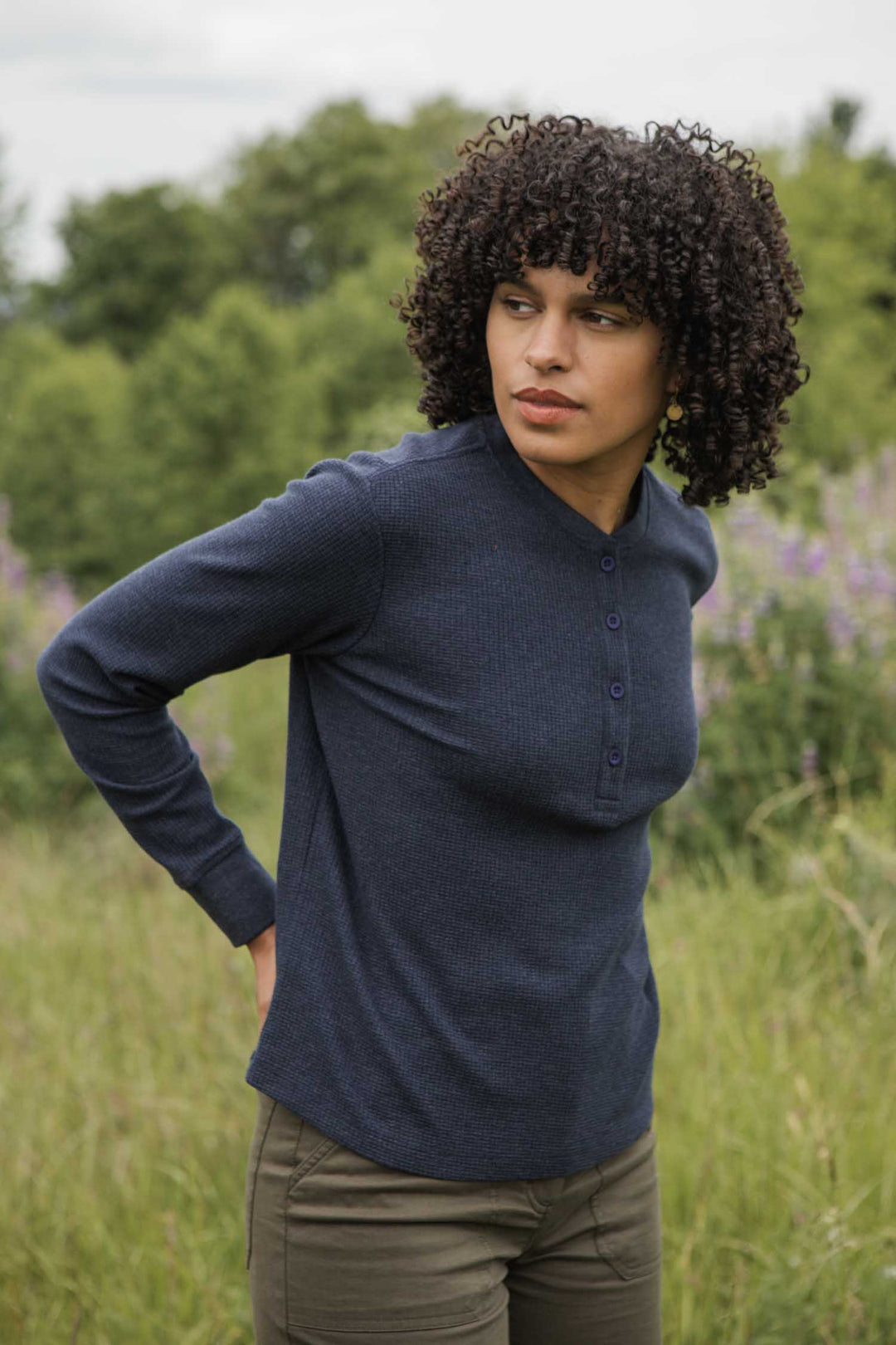 A person with curly hair wears the Bridge & Burn Women's Recycled Waffle Henley in Sapphire Melange and olive pants, standing outdoors in tall grass with trees and greenery behind her.