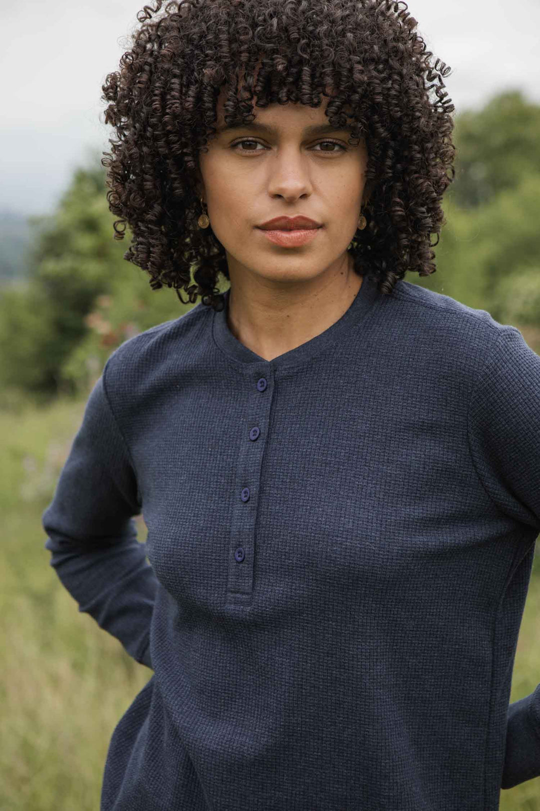 A woman with curly dark hair wears the Bridge & Burn Women's Recycled Waffle Henley in Sapphire Melange, standing in a grassy field with a neutral expression. Trees and greenery create a blurred background outdoors.