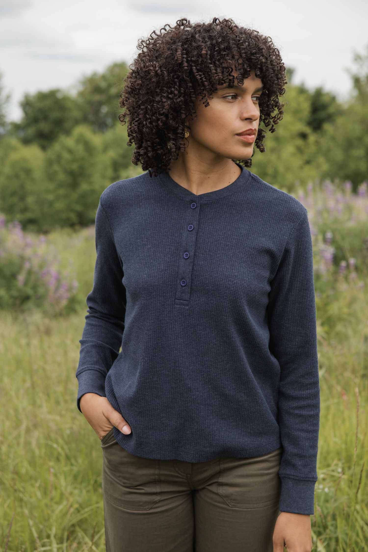 A person with curly hair stands in a grassy field wearing the Bridge & Burn Women's Recycled Waffle Henley in Sapphire Melange and olive green pants, looking to the side with one hand in their pocket among trees and wildflowers.