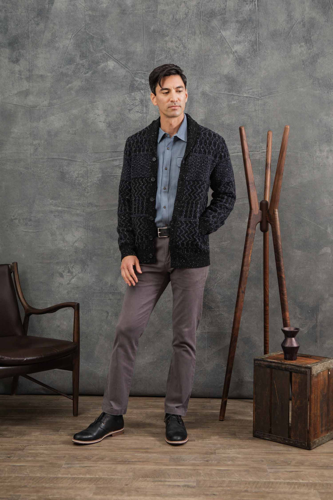 A man stands indoors on wooden flooring wearing the Bridge & Burn Anders Cardigan in Midnight Tweed over a blue shirt, paired with gray pants and black shoes. One hand is in his pocket near a wooden chair, coat rack, and small box.