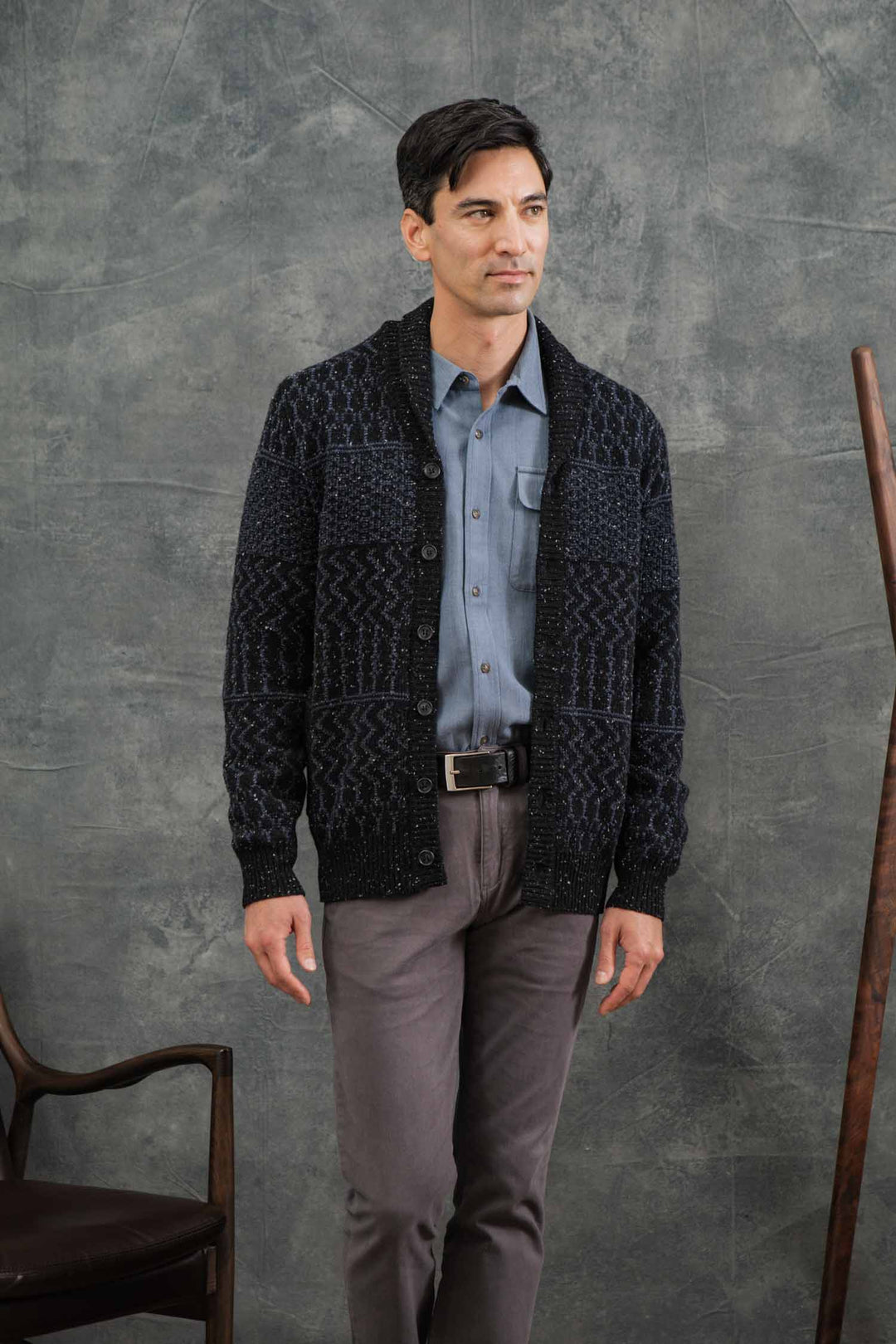 A man with short dark hair stands indoors near a chair and wooden rod, wearing the Bridge & Burn Anders Cardigan in Midnight Tweed (RWS certified wool) over a blue shirt and gray pants, set against a textured gray background.