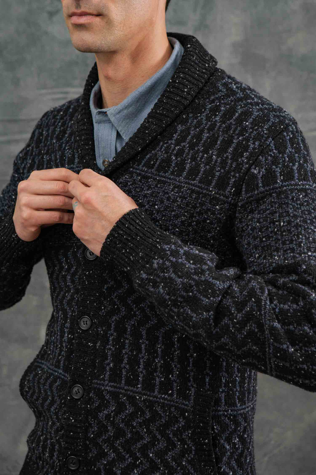 A man, with only the lower half of his face visible, buttons up the Bridge & Burn Anders Cardigan in Midnight Tweed over a light blue collared shirt against a neutral gray background.
