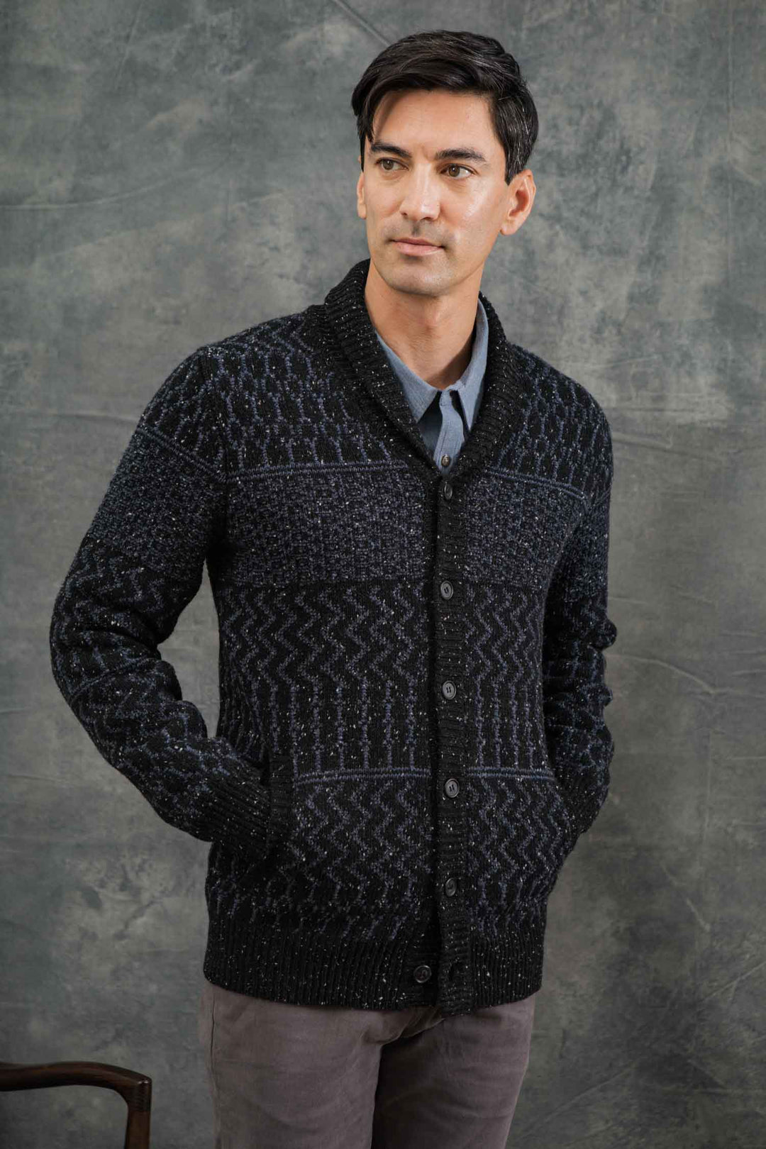 A man stands hands-in-pockets before a textured gray background, wearing the Bridge & Burn Anders Cardigan in Midnight Tweed over a gray collared shirt, looking slightly to the side.