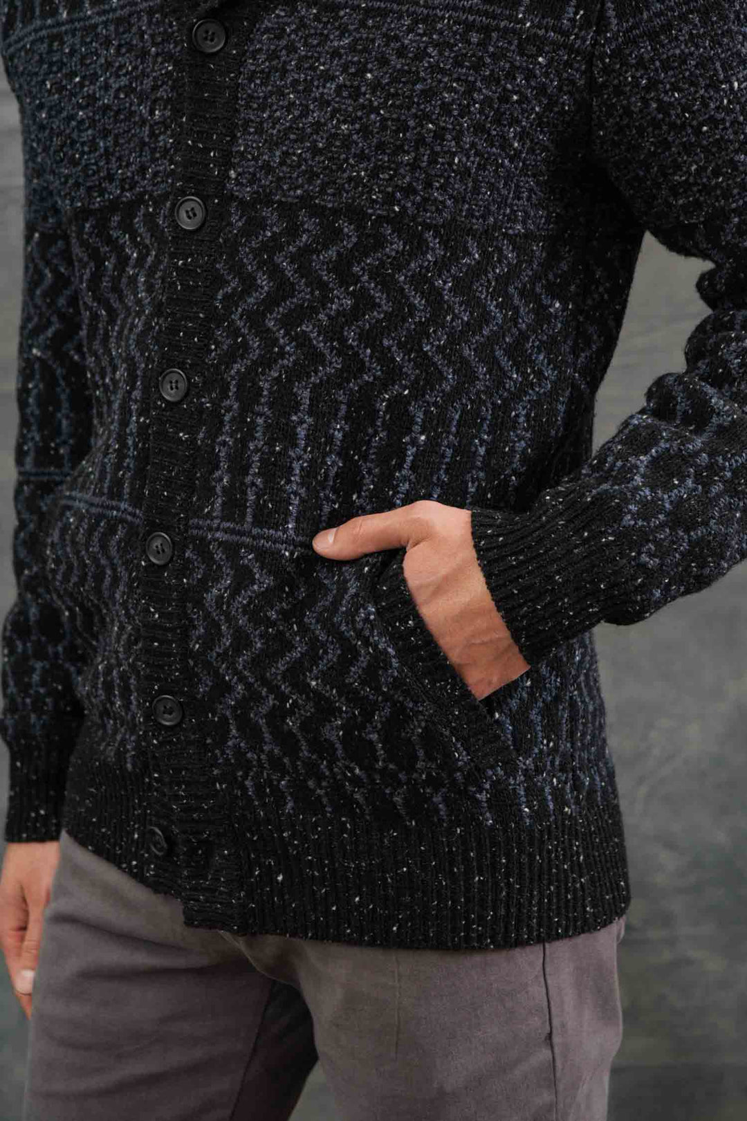 A person wears the Bridge & Burn Anders Cardigan in Midnight Tweed—featuring a textured black and dark gray button-front and cozy shawl collar—with light gray pants, standing against a gray backdrop.