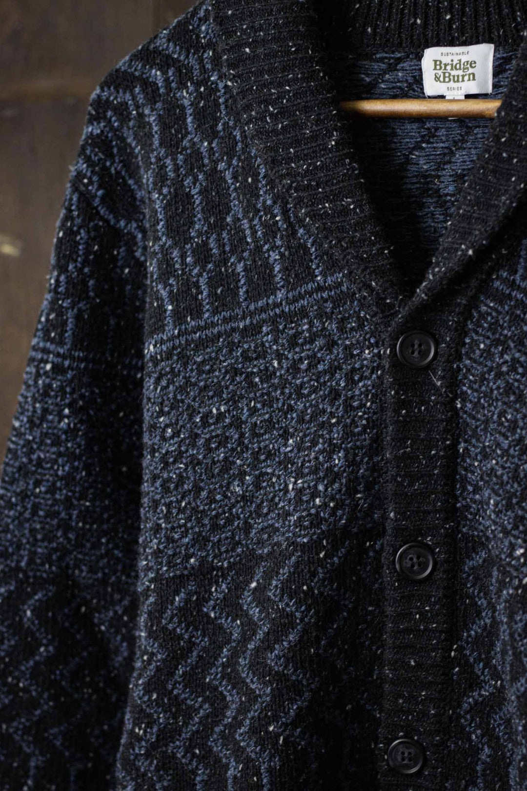 A close-up of the Bridge & Burn Anders Cardigan in Midnight Tweed, featuring dark blue and black textured knit, black buttons, and a cozy shawl collar, hanging on a wooden hanger against a dark background.