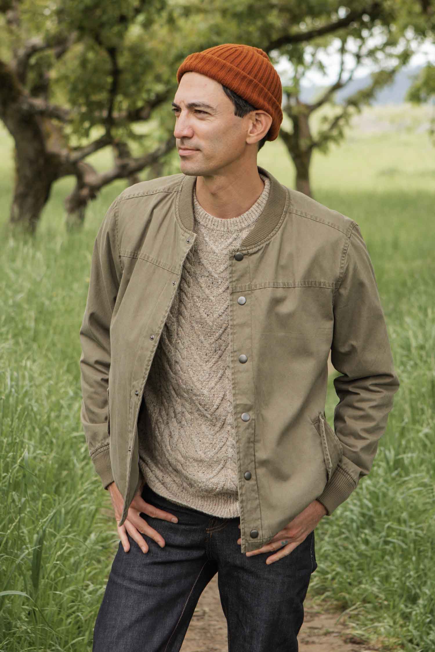 A man wearing a Bridge & Burn Austen Varsity Jacket in khaki green canvas stands outdoors in a grassy field with trees in the background, looking to the side with his hands in his jacket pockets.