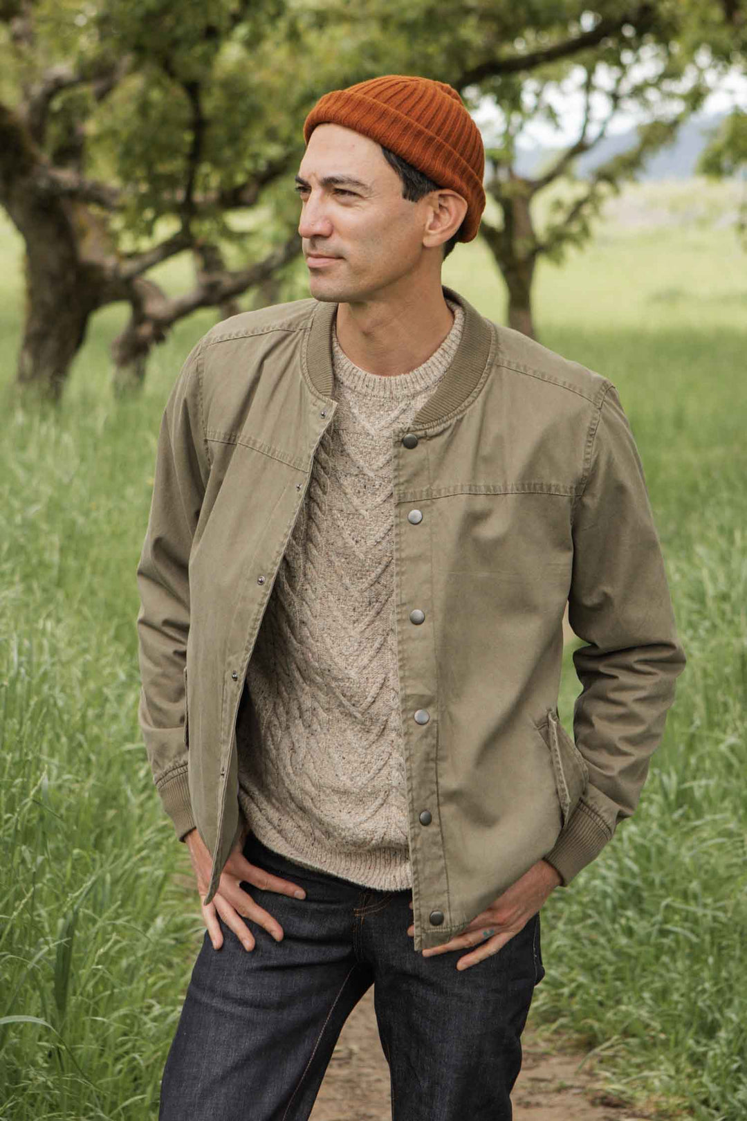 A man wearing a Bridge & Burn Austen Varsity Jacket in khaki green canvas stands outdoors in a grassy field with trees in the background, looking to the side with his hands in his jacket pockets.