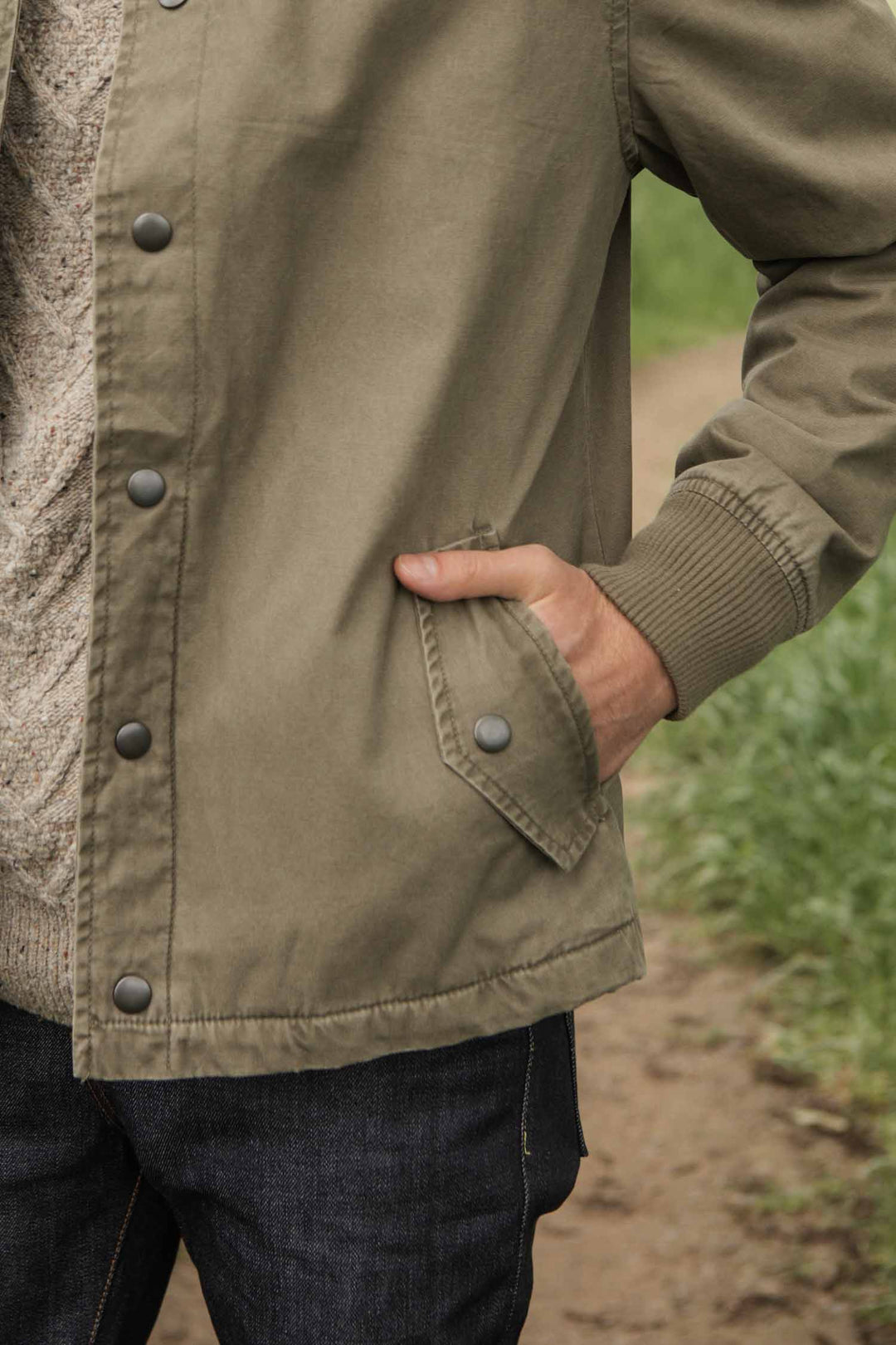 A person wears the Bridge & Burn Austen Varsity Jacket in Khaki Green Canvas and dark jeans outdoors, with one hand in their jacket pocket; only their torso and part of one arm are visible.