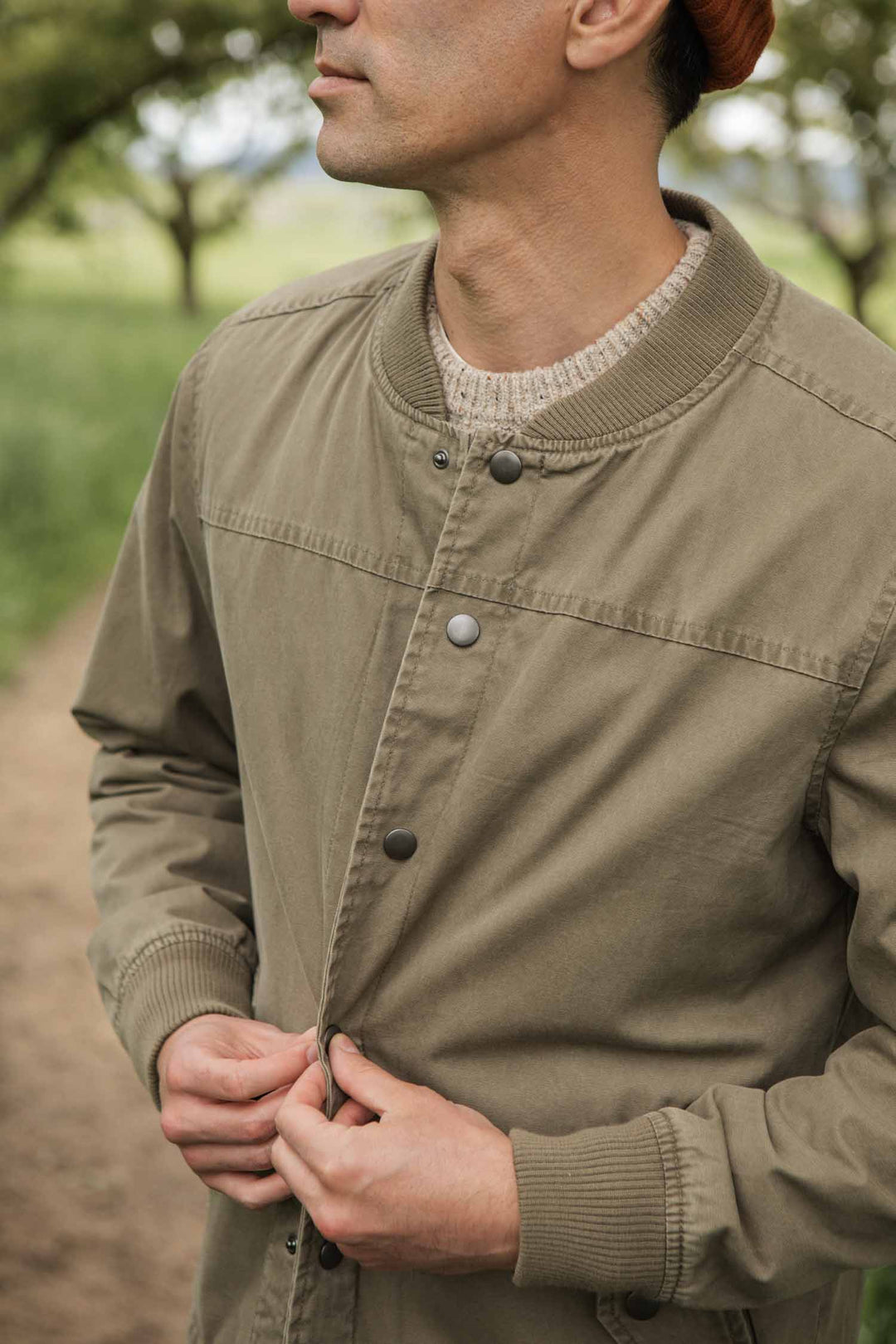 A man outdoors buttons up the Bridge & Burn Austen Varsity Jacket in khaki green canvas over a beige sweater. His face is partially visible, with leafy trees blurred in the background.