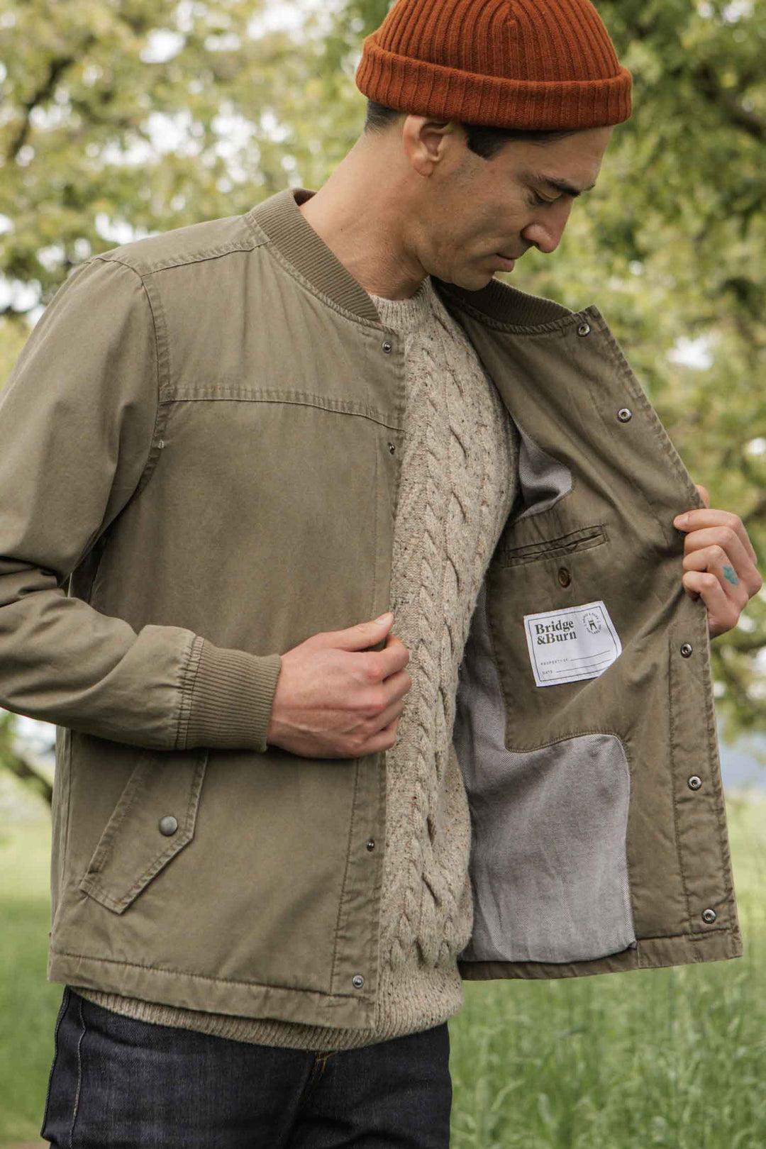 A man outdoors wears the Bridge & Burn Austen Varsity Jacket in Khaki Green Canvas, paired with a beige cable-knit sweater and an orange knit beanie. He opens the jacket to display its inner label, standing before blurred greenery.
