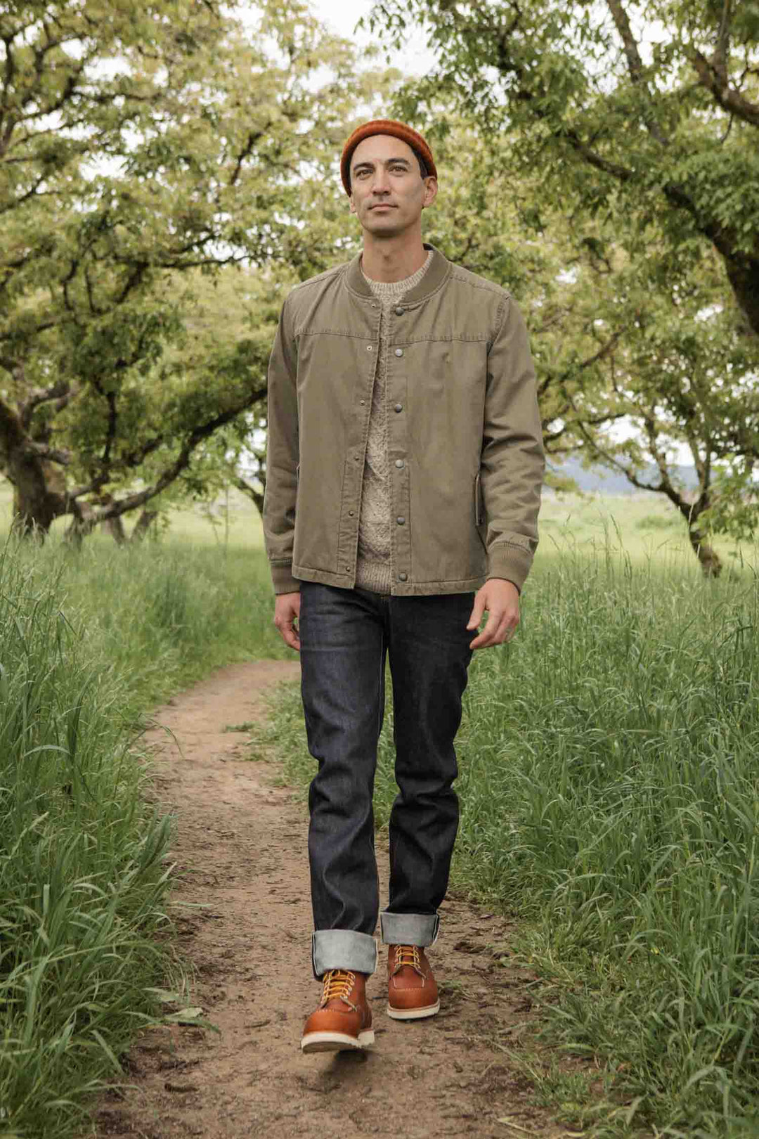 A man wearing the Bridge & Burn Austen Varsity Jacket in Khaki Green Canvas, an orange beanie, jeans, and brown boots walks along a dirt path through tall grass and trees outdoors.
