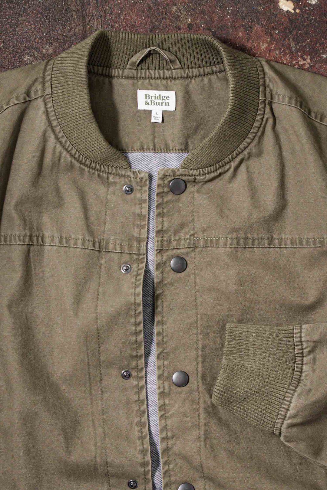 A close-up of the Bridge & Burn Austen Varsity Jacket in khaki green canvas, featuring a ribbed collar and cuffs, snap button closures, and a front pocket.