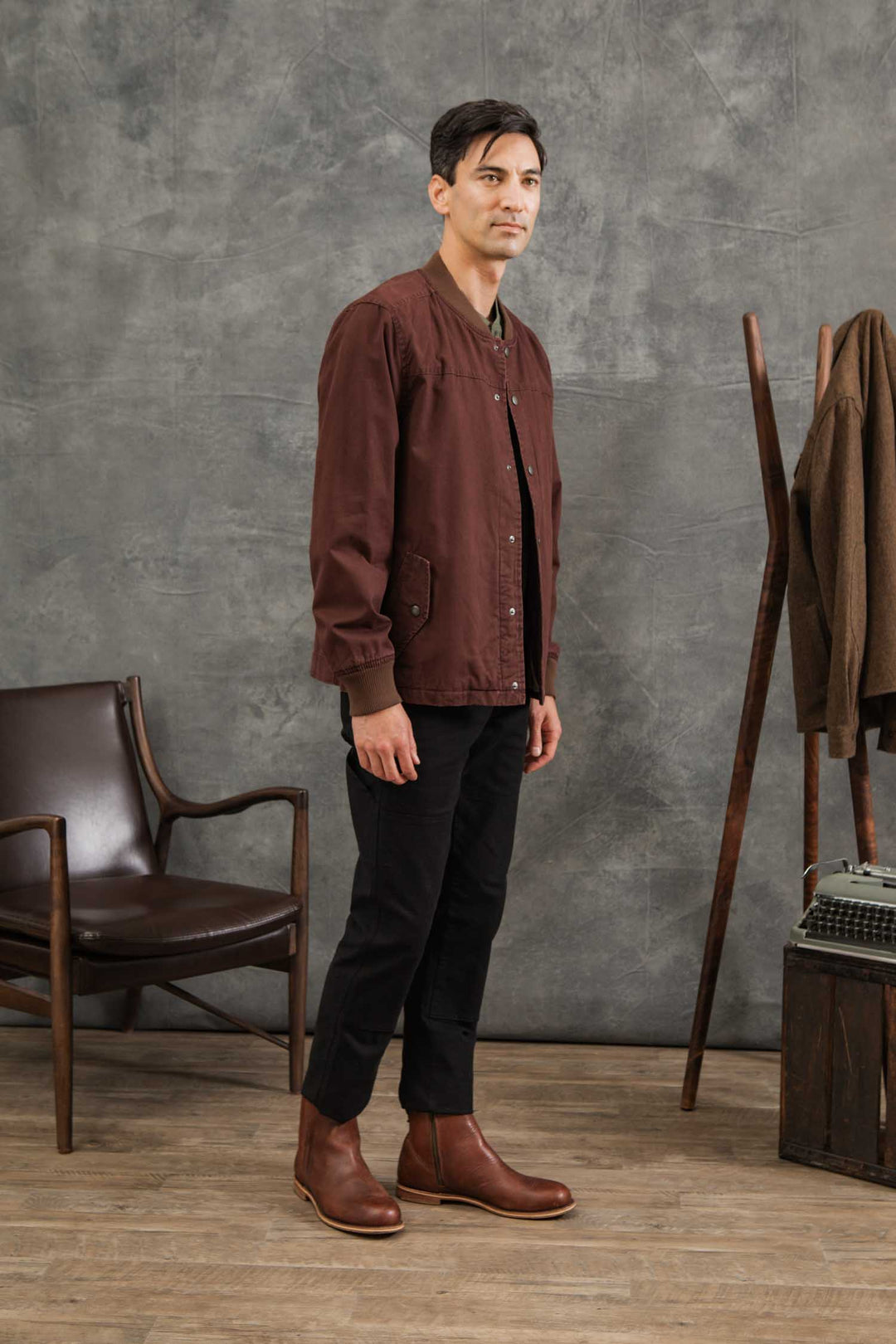 A man stands indoors next to a brown chair and coat rack, wearing the Bridge & Burn Austen Varsity Jacket in Mahogany Canvas, black pants, and brown boots against a gray textured backdrop.