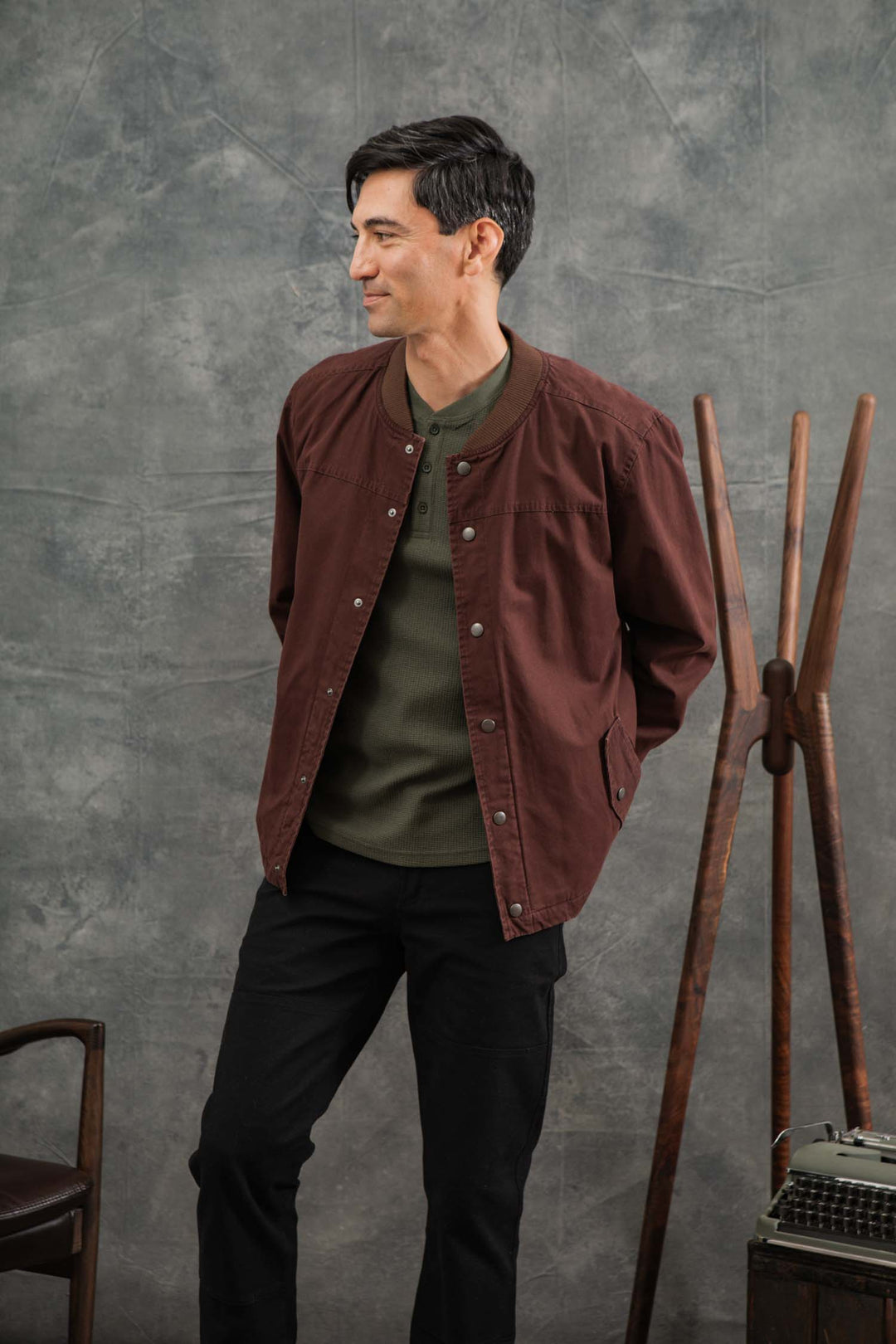 A man with short dark hair stands before a gray textured backdrop, hands in his pockets, wearing the Bridge & Burn Austen Varsity Jacket in Mahogany Canvas over a green shirt and black pants. He looks to his left with a slight smile.