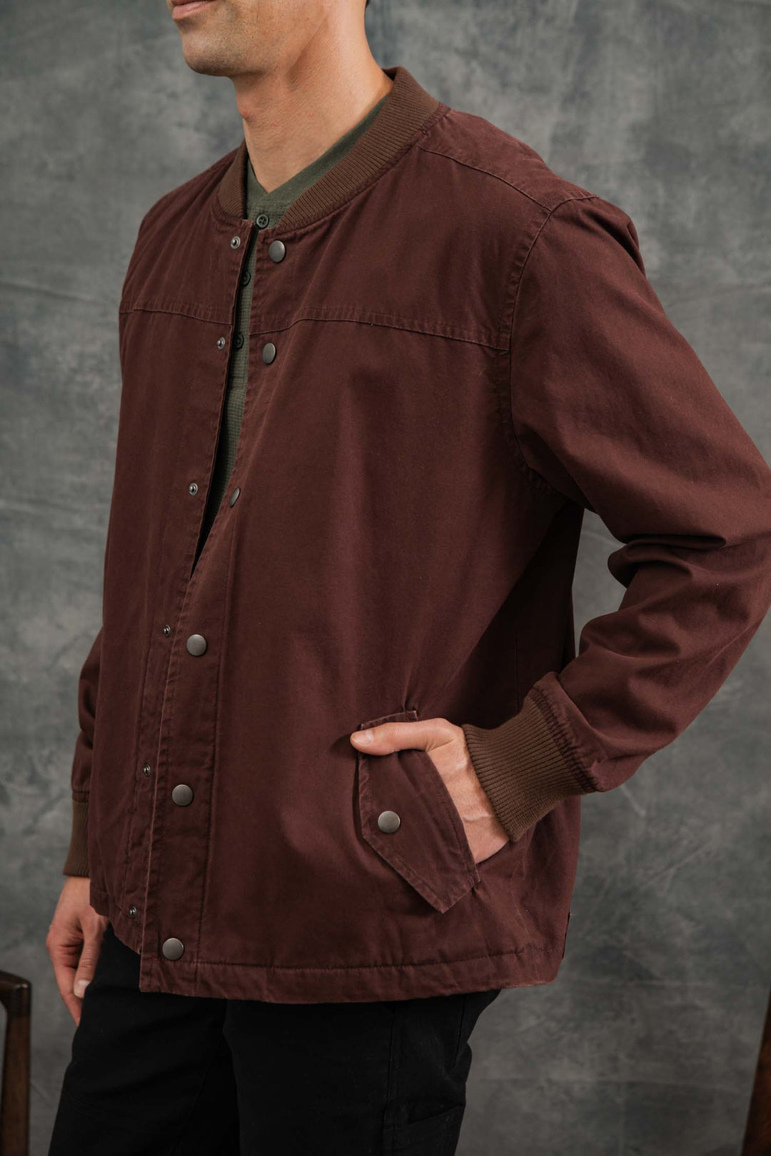 A man stands with one hand in his pocket against a gray textured background, modeling the Bridge & Burn Austen Varsity Jacket in Mahogany Canvas. His face is partially out of frame.
