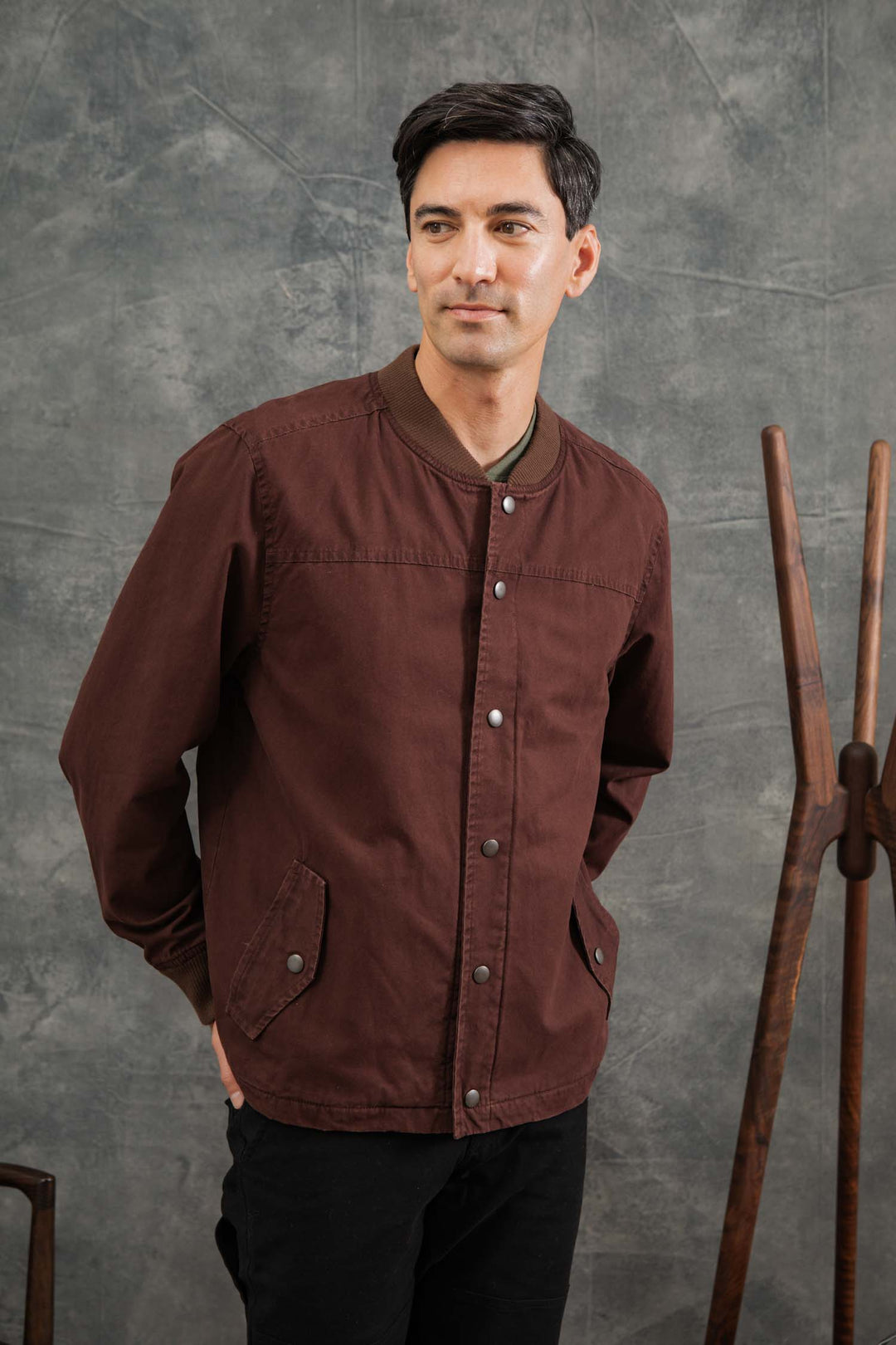 A man with short dark hair wears the Bridge & Burn Austen Varsity Jacket in Mahogany Canvas over a brown shirt, standing with hands in pockets before a textured gray backdrop and wooden coat rack.