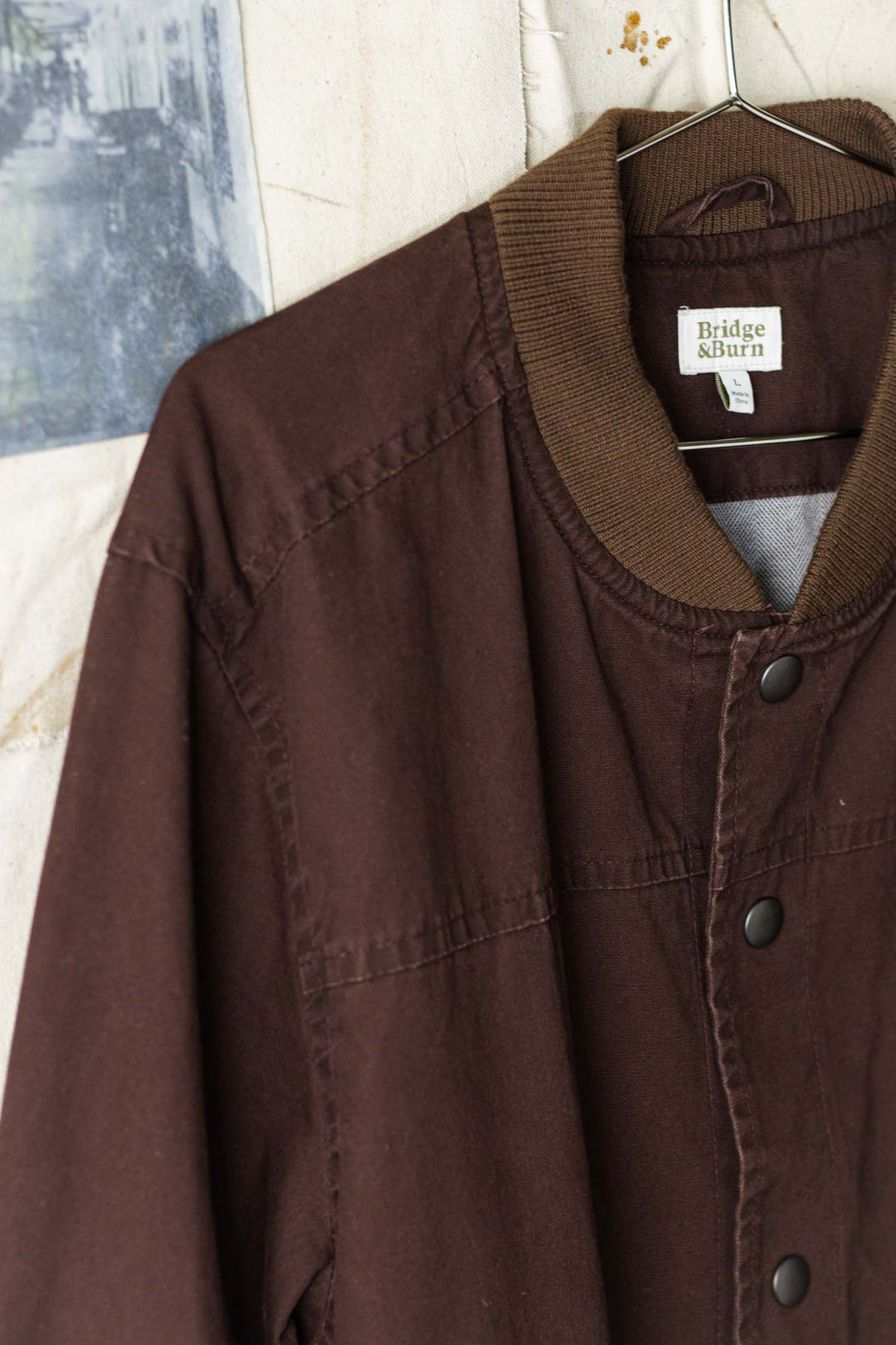 The Austen Varsity Jacket in mahogany canvas by Bridge & Burn hangs on a wire hanger, featuring a ribbed collar and snap buttons. Its cotton shell offers comfort, with a blurred bluish background image partially visible.