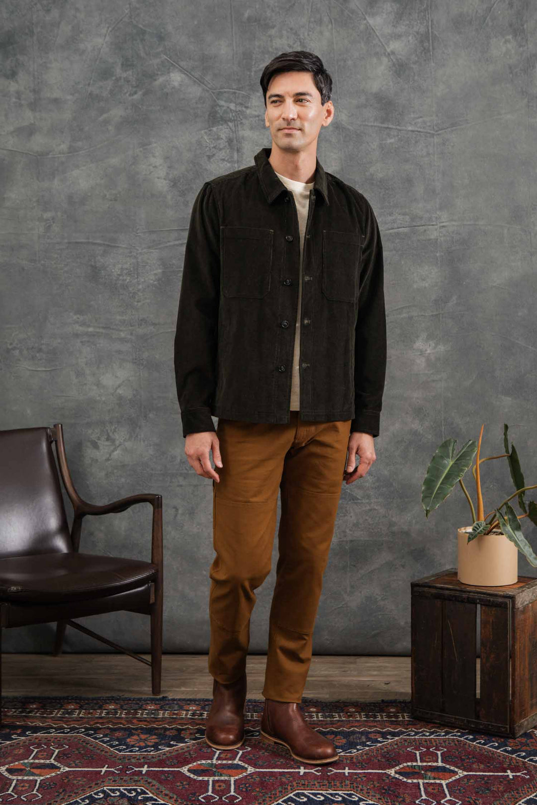A man stands indoors on a patterned rug, wearing the Bridge & Burn Banks Shirt Jacket in Deep Pine Corduroy over a beige shirt with brown pants and boots. He is beside a brown chair and small plant, set against a gray textured wall.