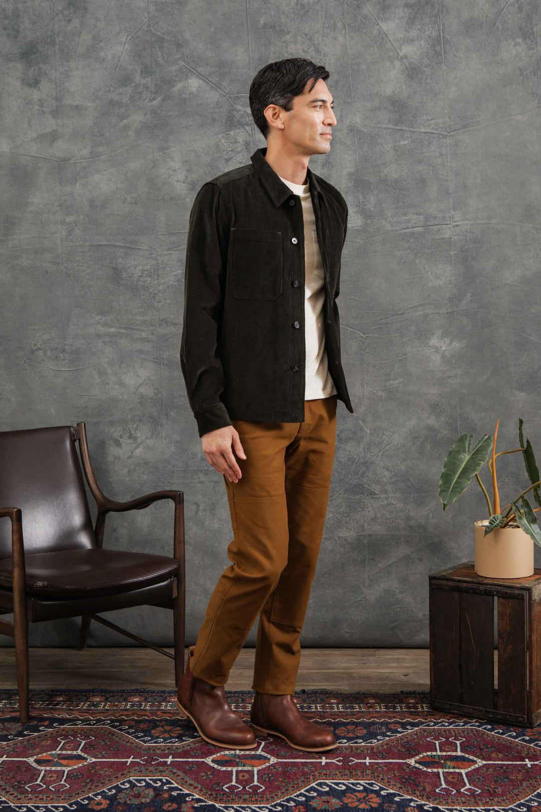 A man stands indoors on a patterned rug, wearing the Bridge & Burn Banks Shirt Jacket in Deep Pine Corduroy. His classic workwear-inspired look is complemented by a leather chair, wooden chest, and potted plant nearby.