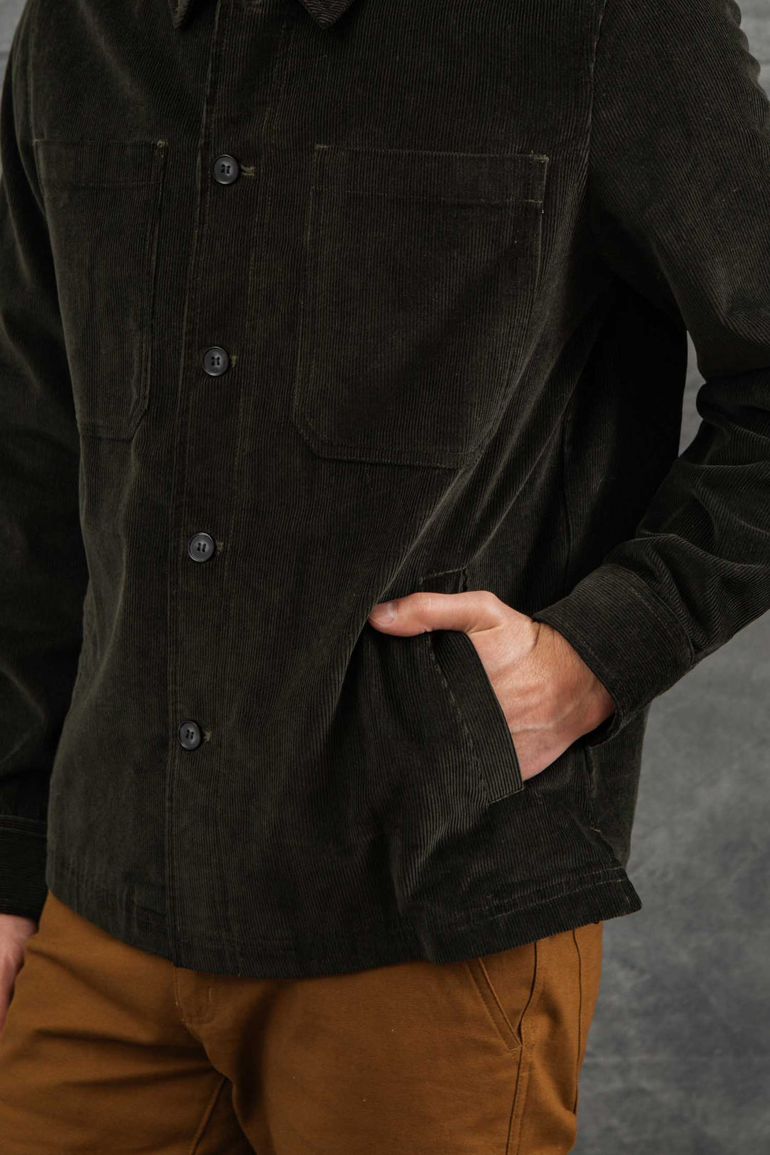 A person wearing the Bridge & Burn Banks Shirt Jacket in Deep Pine Corduroy with front pockets and brown pants stands with one hand in their pocket against a gray textured background.