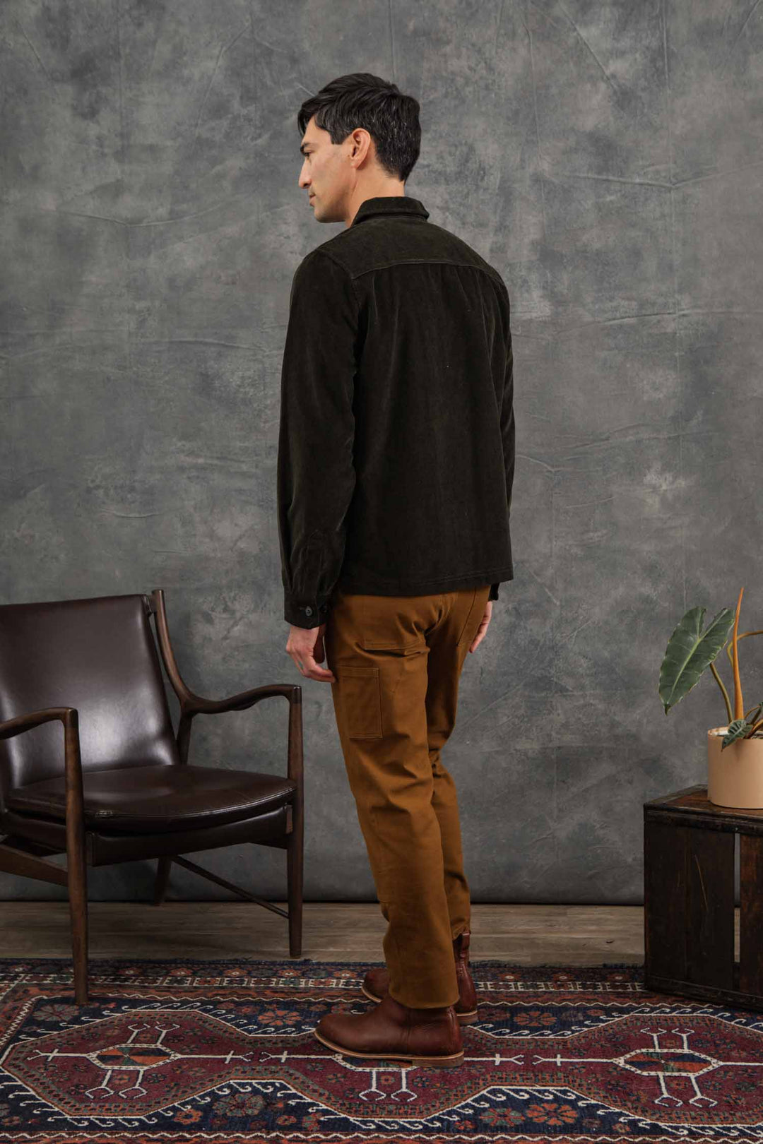 A man stands indoors facing sideways, wearing the Bridge & Burn Banks Shirt Jacket in Deep Pine Corduroy and tan pants. He is complemented by a brown leather chair, patterned rug, potted plant, and a gray textured wall in the background.