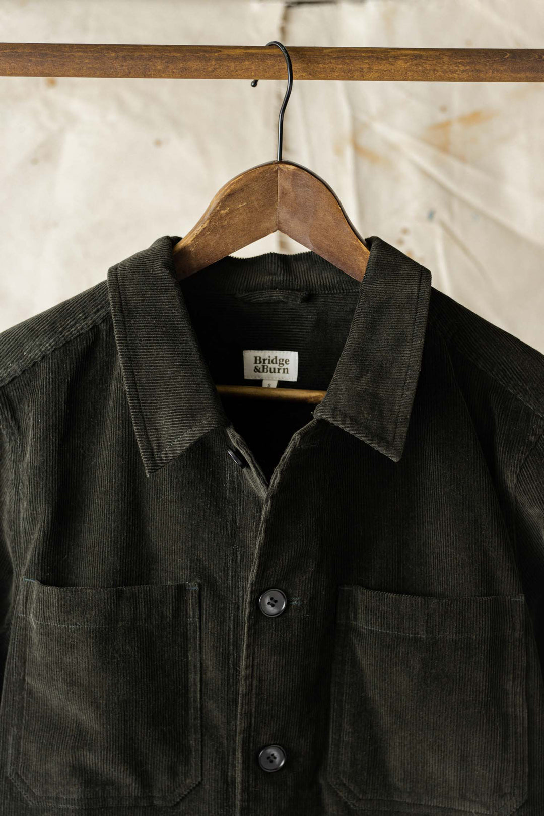 The Bridge & Burn Banks Shirt Jacket in deep pine corduroy, featuring large buttons and front pockets, hangs on a wooden hanger against a light textured background.