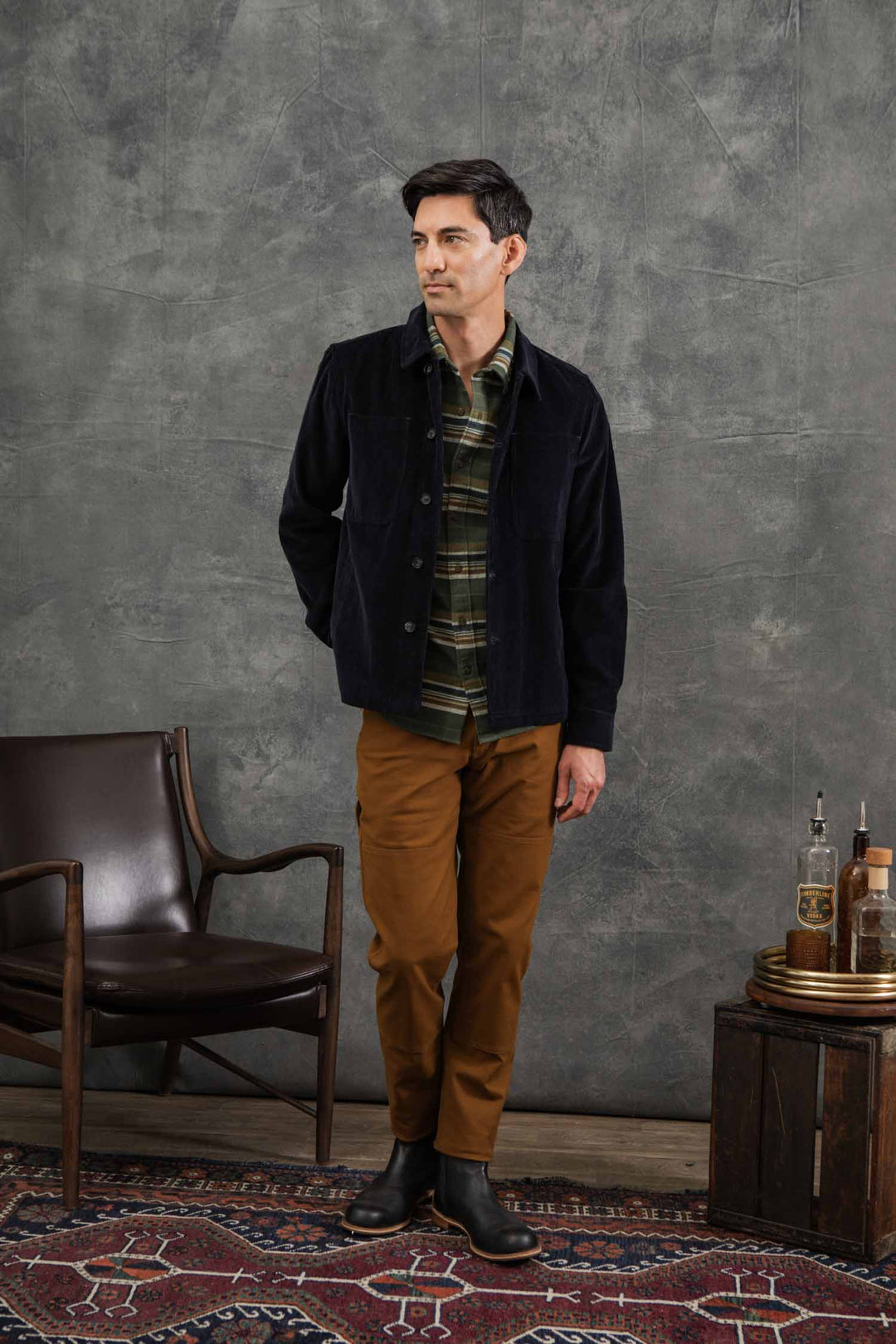 A man stands indoors on a patterned rug, wearing the Bridge & Burn Banks Shirt Jacket in Midnight Blue Corduroy over a green plaid shirt, tan pants, and black boots. He is beside a brown chair and small wooden table with bottles and glasses, against a gray wall.