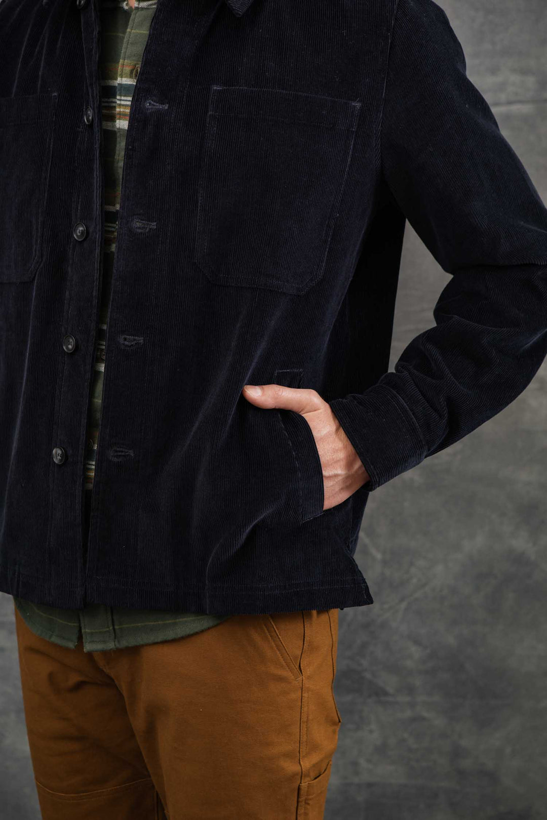 A person wearing the Bridge & Burn Banks Shirt Jacket in Midnight Blue Corduroy over a green plaid shirt and brown pants stands with one hand in their jacket pocket against a gray textured background.
