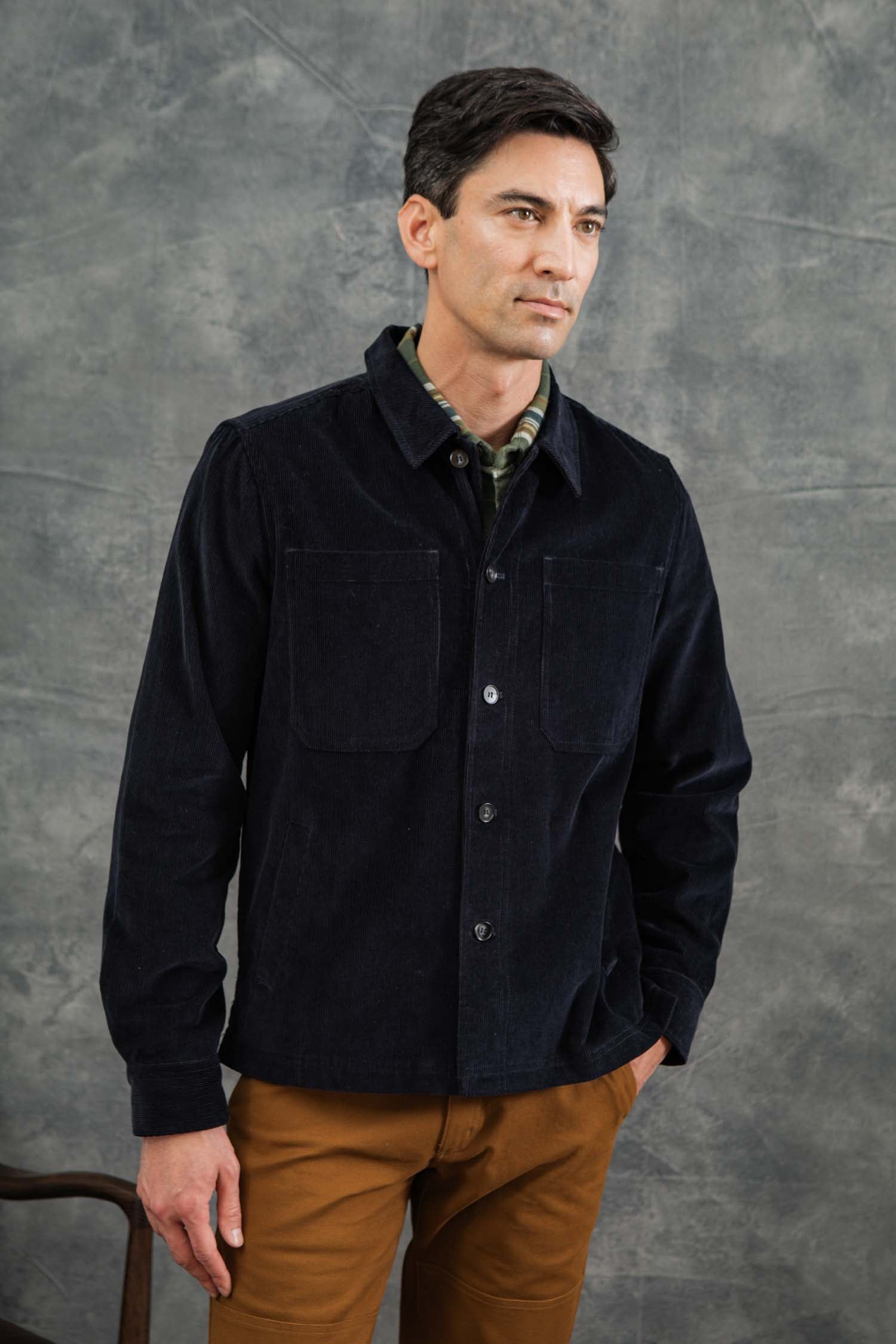 A man with short dark hair wears the Bridge & Burn Banks Shirt Jacket in Midnight Blue Corduroy over a green plaid shirt and brown pants, standing before a textured gray backdrop with a neutral expression, evoking classic workwear style.