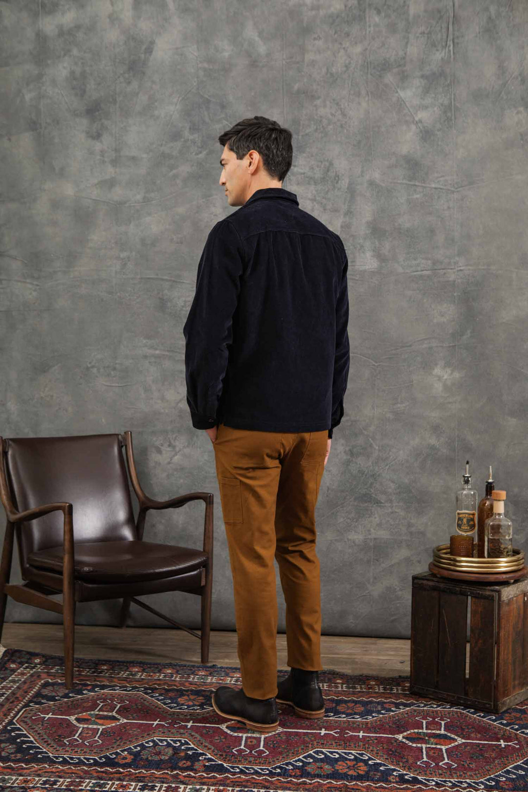 A man with short dark hair stands facing away, wearing the Bridge & Burn Banks Shirt Jacket in midnight blue corduroy and brown pants. He is beside a brown leather chair and small table with bottles, on a rug against a textured gray wall.