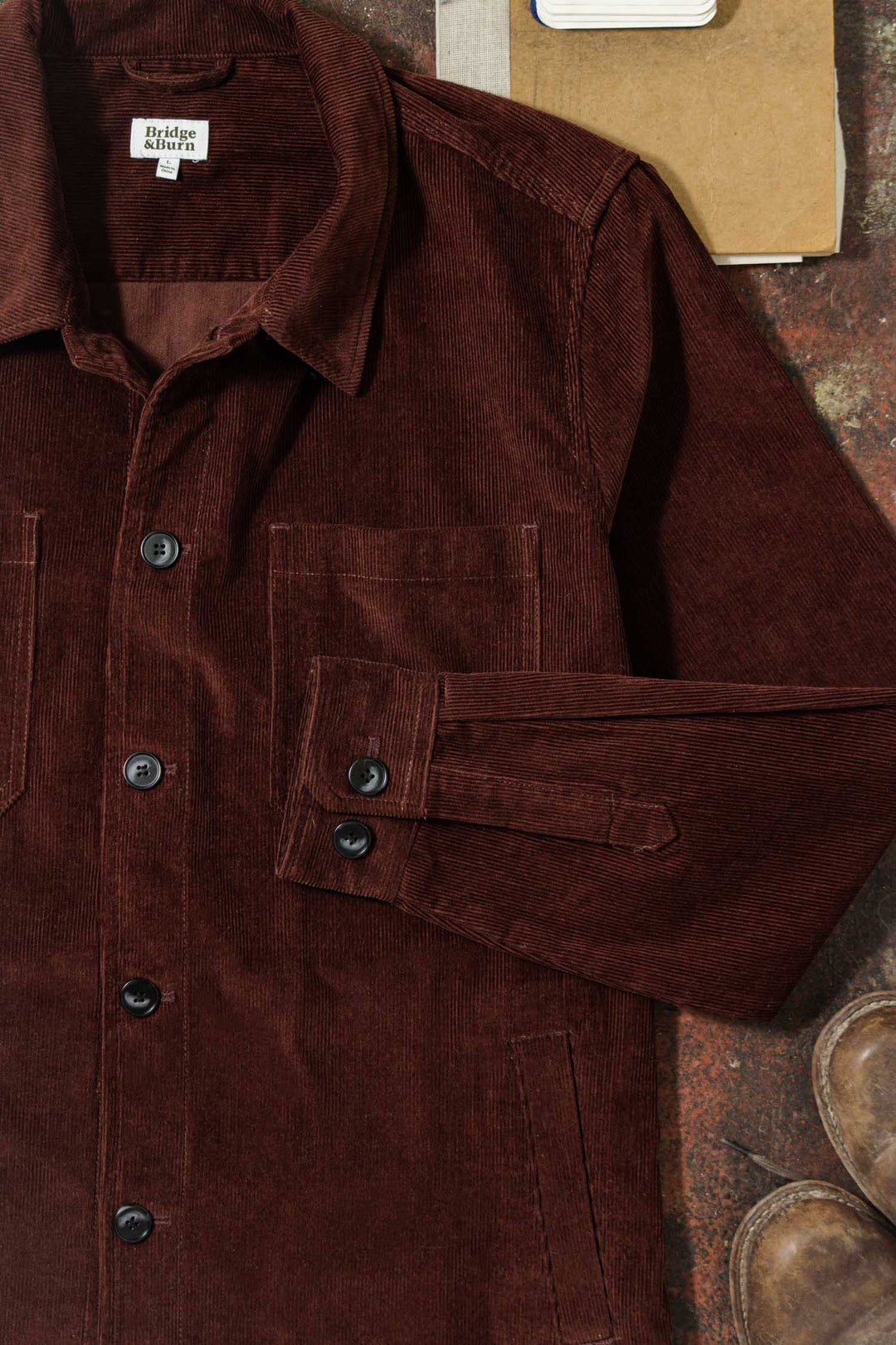 The Bridge & Burn Banks Shirt Jacket in Redwood Corduroy, featuring black buttons, a collared neckline, two front pockets, and a folded left sleeve, is shown laid flat with part of a boot and notebook for a workwear-inspired look.