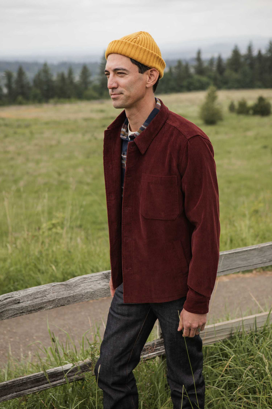 A man in a yellow beanie, Bridge & Burn Banks Shirt Jacket in Redwood Corduroy, and dark jeans stands outdoors by a wooden fence with green grass and trees behind him.