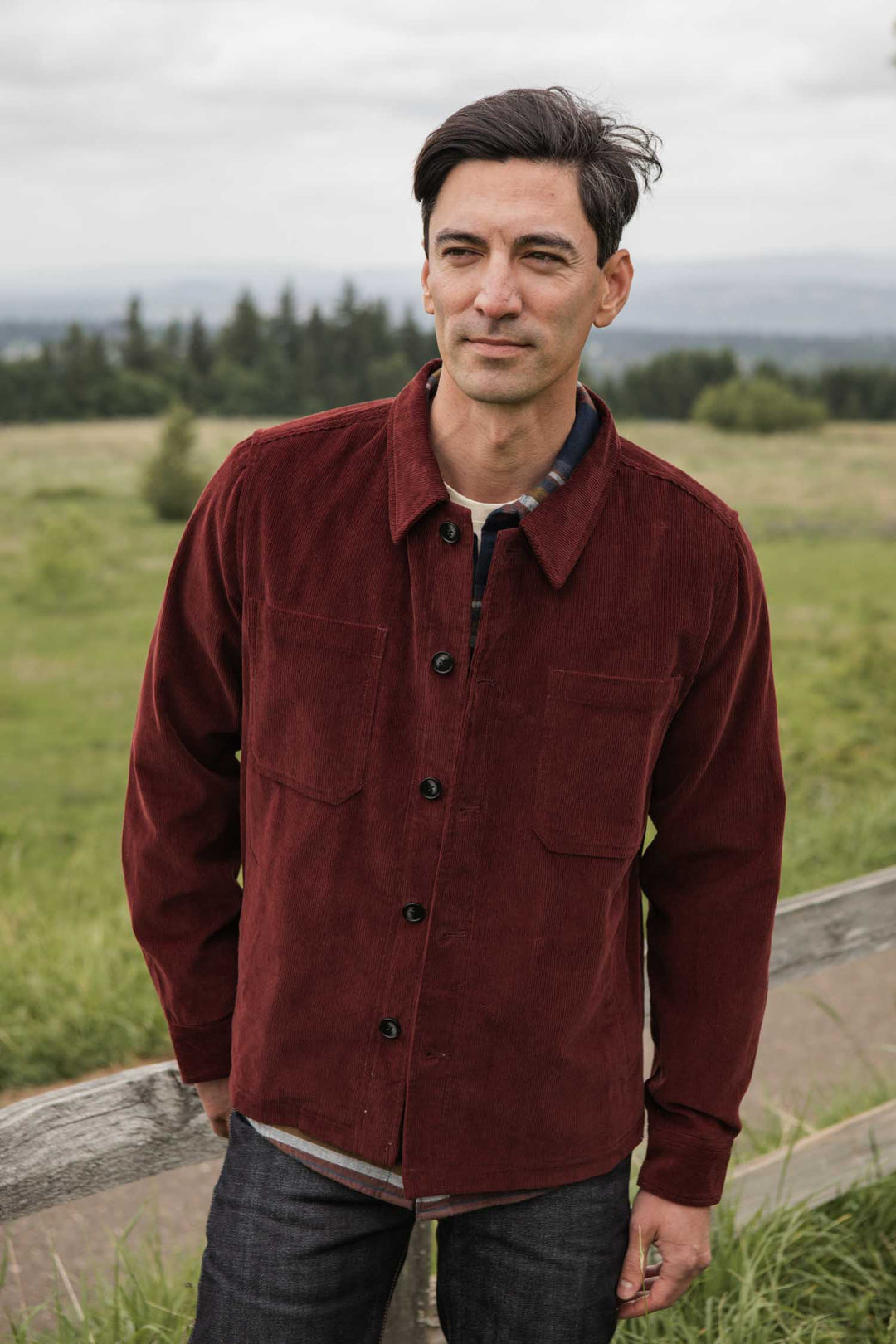 A man wearing the Bridge & Burn Banks Shirt Jacket in Redwood Corduroy stands outdoors by a grassy field and wooden fence, with trees and a cloudy sky behind him.