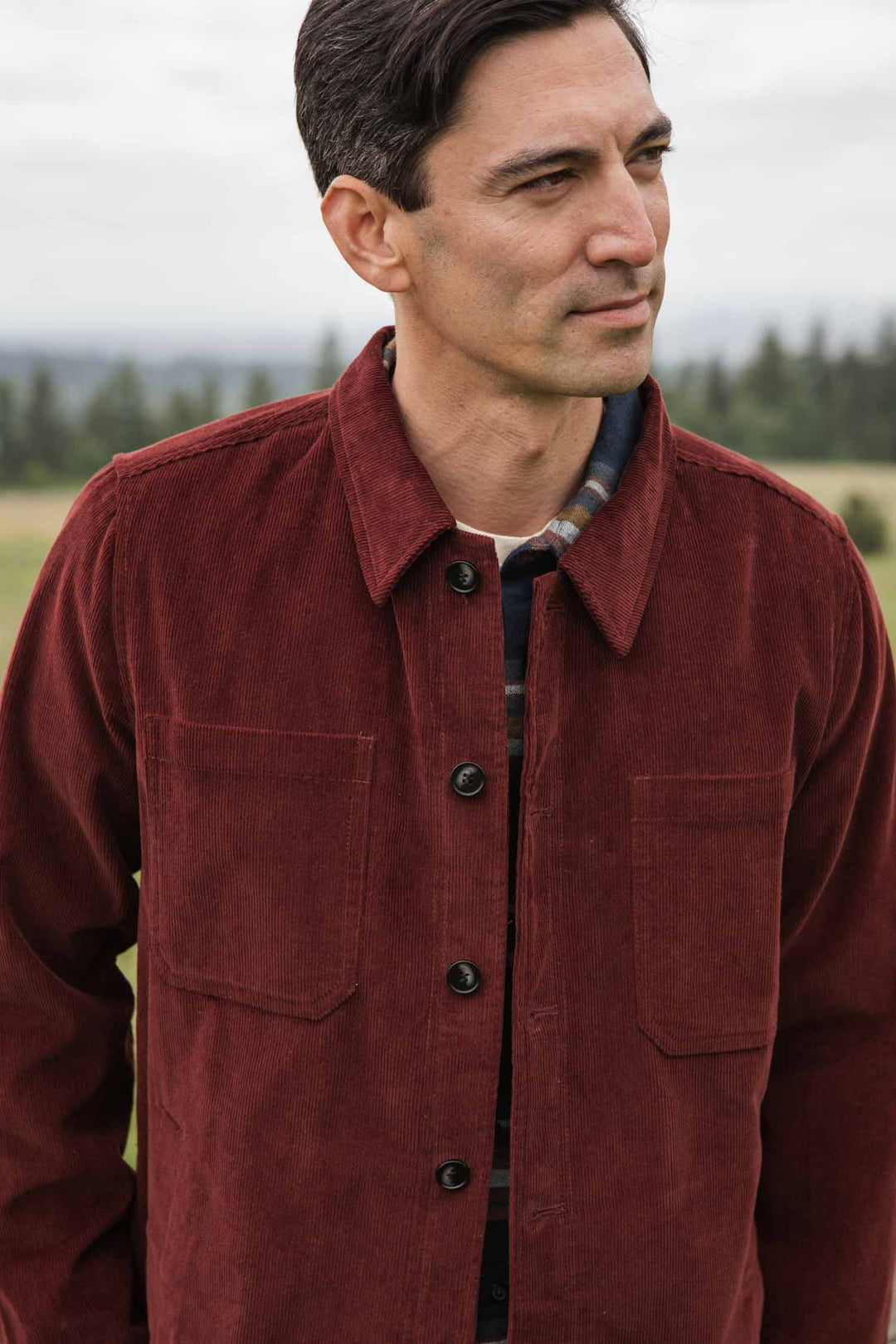 A man wears the Bridge & Burn Banks Shirt Jacket in Redwood Corduroy over a plaid shirt, standing outdoors and smiling slightly. Blurred greenery and trees in the background highlight his relaxed, workwear-inspired look.