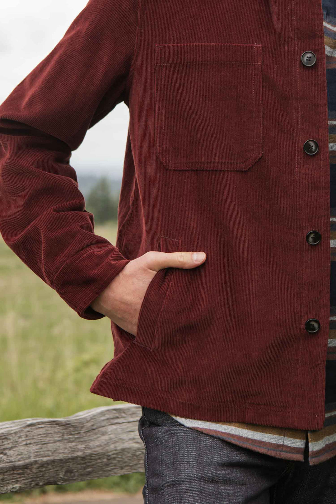 Wearing the Bridge & Burn Banks Shirt Jacket in Redwood Corduroy over a striped shirt, a person stands outdoors by a wooden fence with a hand in their pocket, showcasing laidback workwear style. Only the torso and part of an arm are visible.