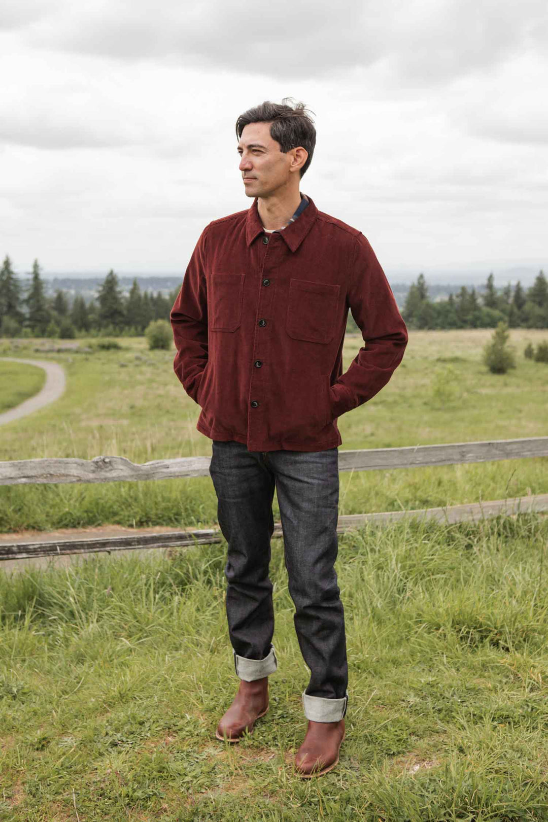 A man stands on grass before a wooden fence, wearing the Bridge & Burn Banks Shirt Jacket in Redwood Corduroy, dark cuffed jeans, and brown boots. Trees and a cloudy sky are visible in the background.