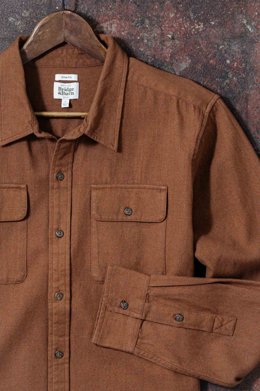 The Bridge & Burn Bedford Shirt in Terracotta Melange, made from brown organic cotton and featuring two chest pockets, hangs on a wooden hanger against a textured dark red and brown background.