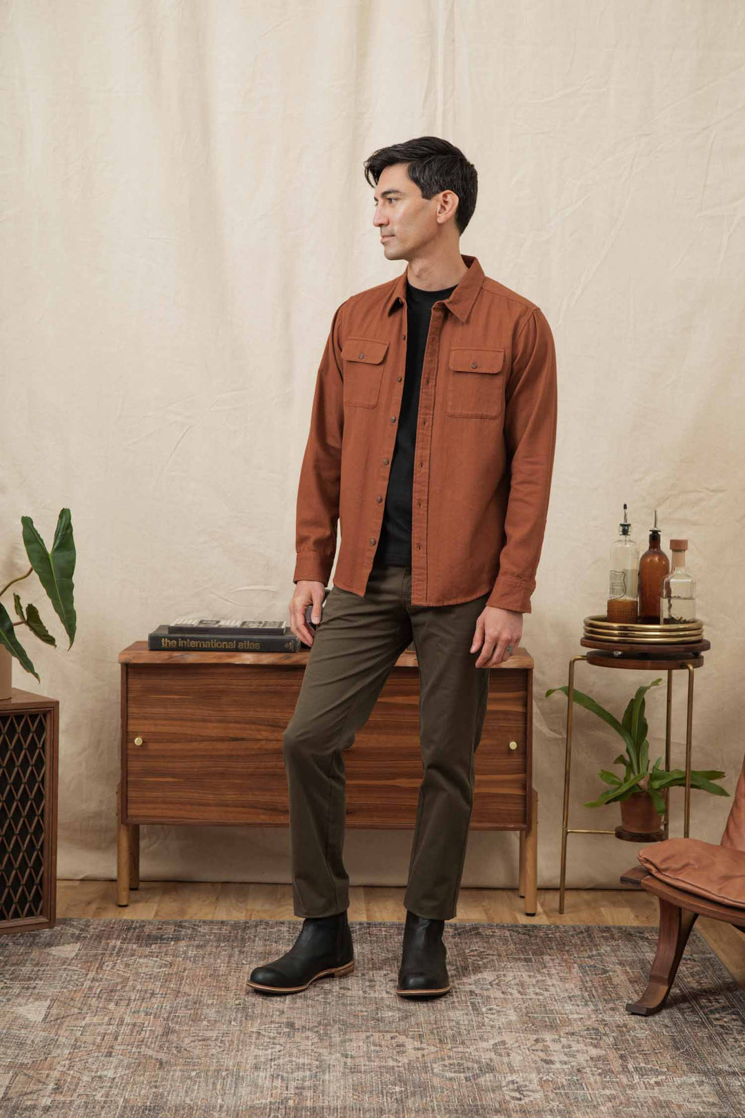 A man stands indoors on a patterned rug, wearing the Bridge & Burn Bedford Shirt in Terracotta Melange over a black t-shirt, olive green pants, and black boots. He faces left, with mid-century modern furniture and plants behind him.