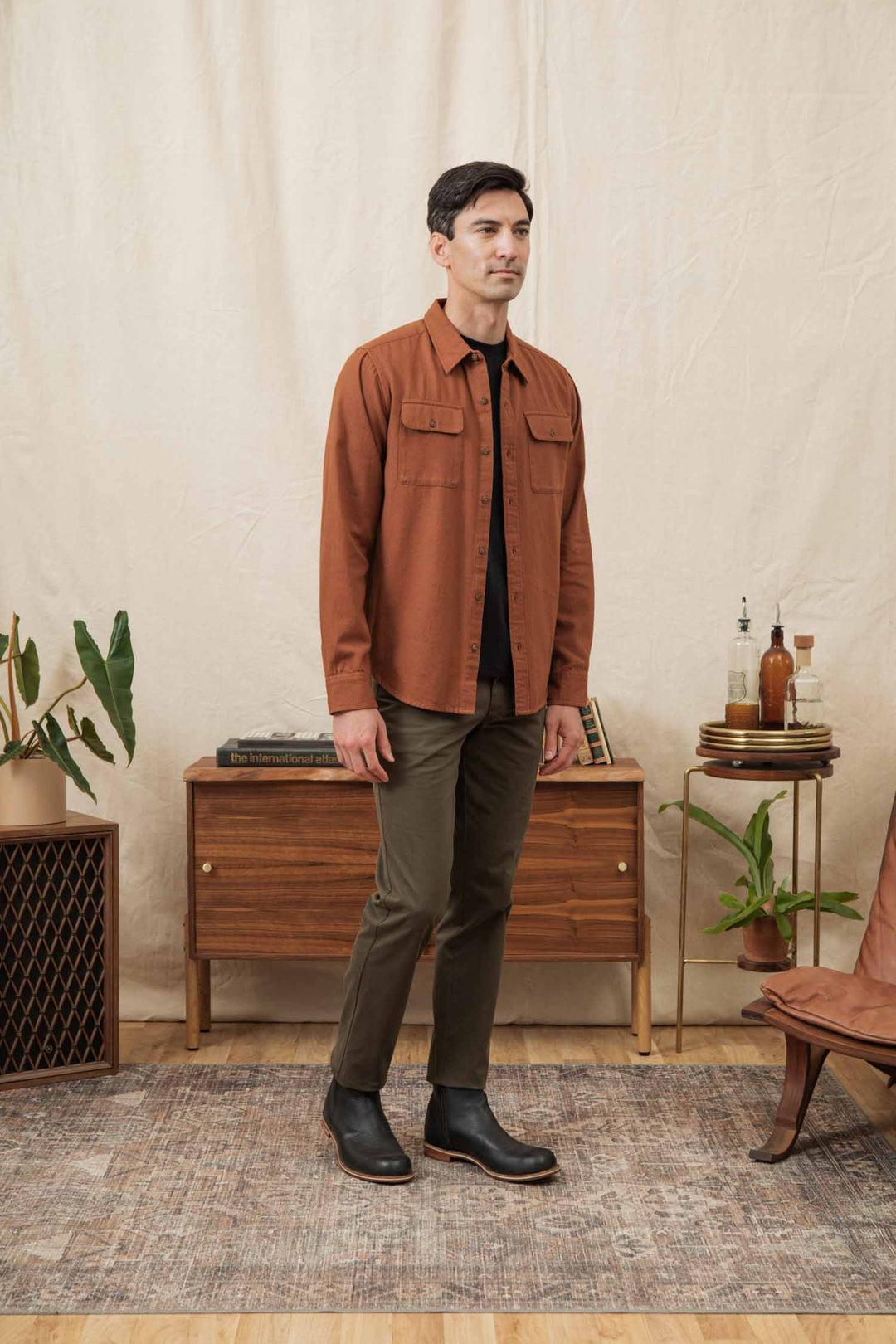 A man stands indoors on a patterned rug, wearing the Bridge & Burn Bedford Shirt in Terracotta Melange, dark olive pants, and black boots. Behind him are a wooden sideboard with decor and several potted plants.