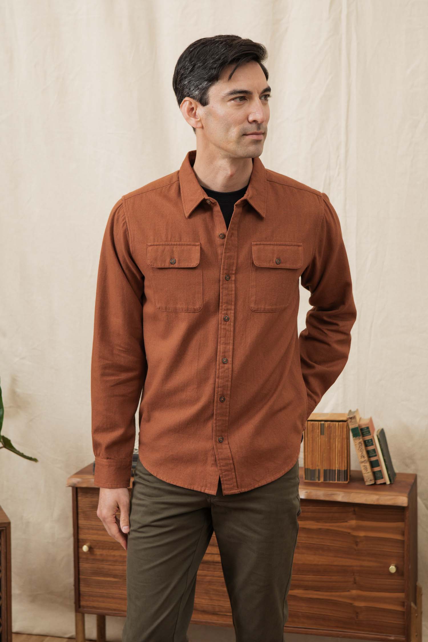 A man with short dark hair wears the Bridge & Burn Bedford Shirt in Terracotta Melange and olive green pants, standing indoors before a light-colored curtain and a wooden table topped with stacked books.