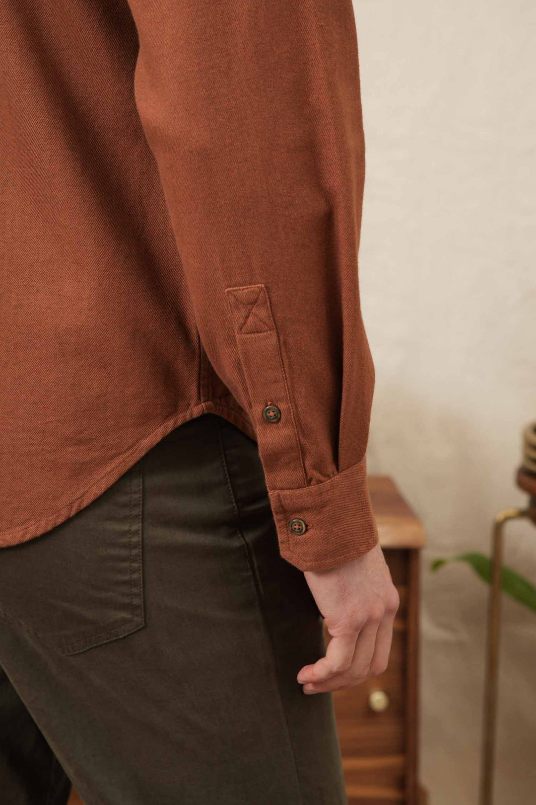 A close-up of a person’s arm and hand wearing the Bridge & Burn Bedford Shirt in Terracotta Melange with dark olive-green pants. A small wooden table and a plant appear in the background.