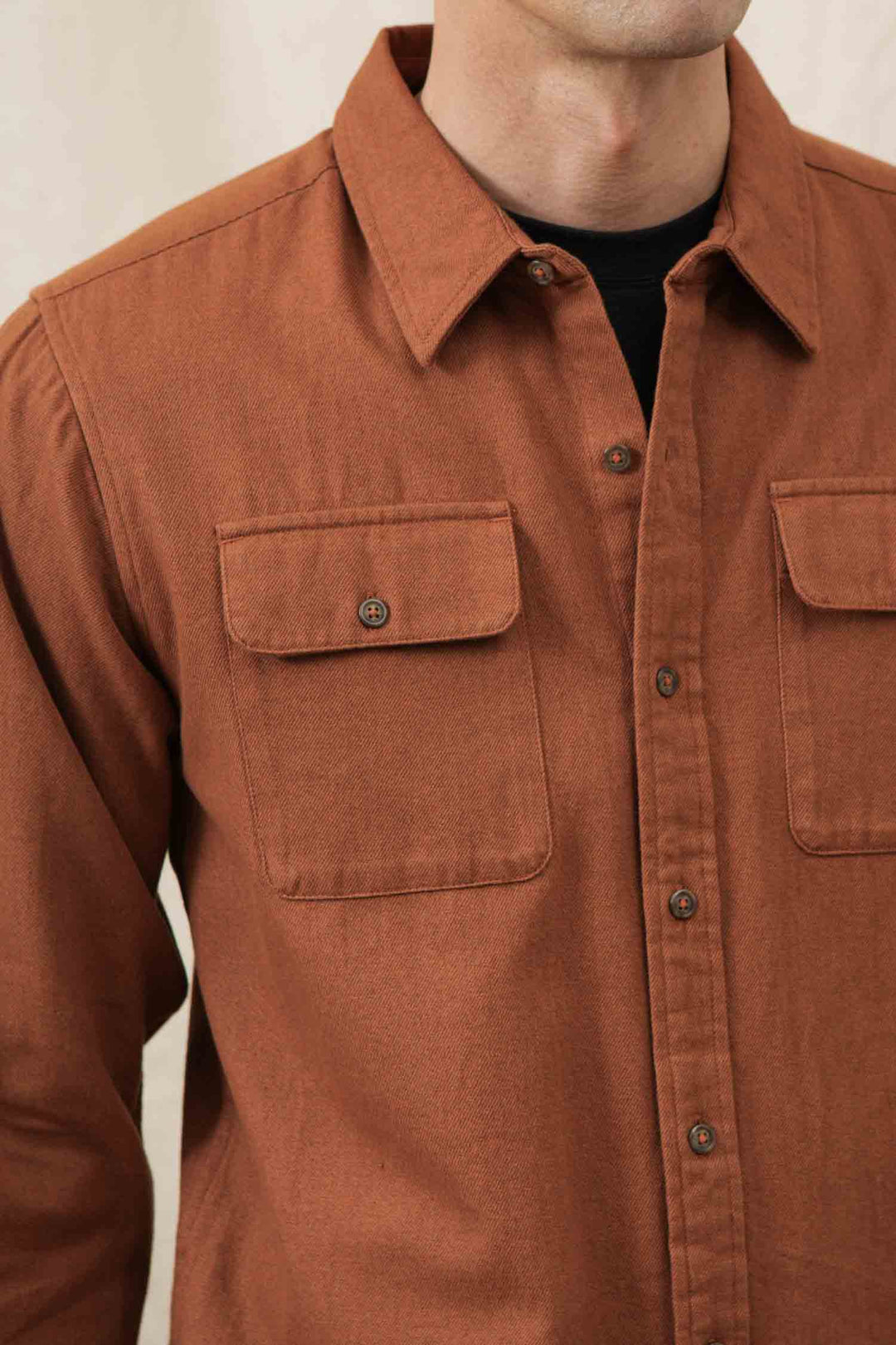 A person models the Bridge & Burn Bedford Shirt in Terracotta Melange—an organic cotton overshirt with two chest pockets—unbuttoned at the top to reveal a black shirt underneath, while standing against a light background. The person's head is slightly out of frame.