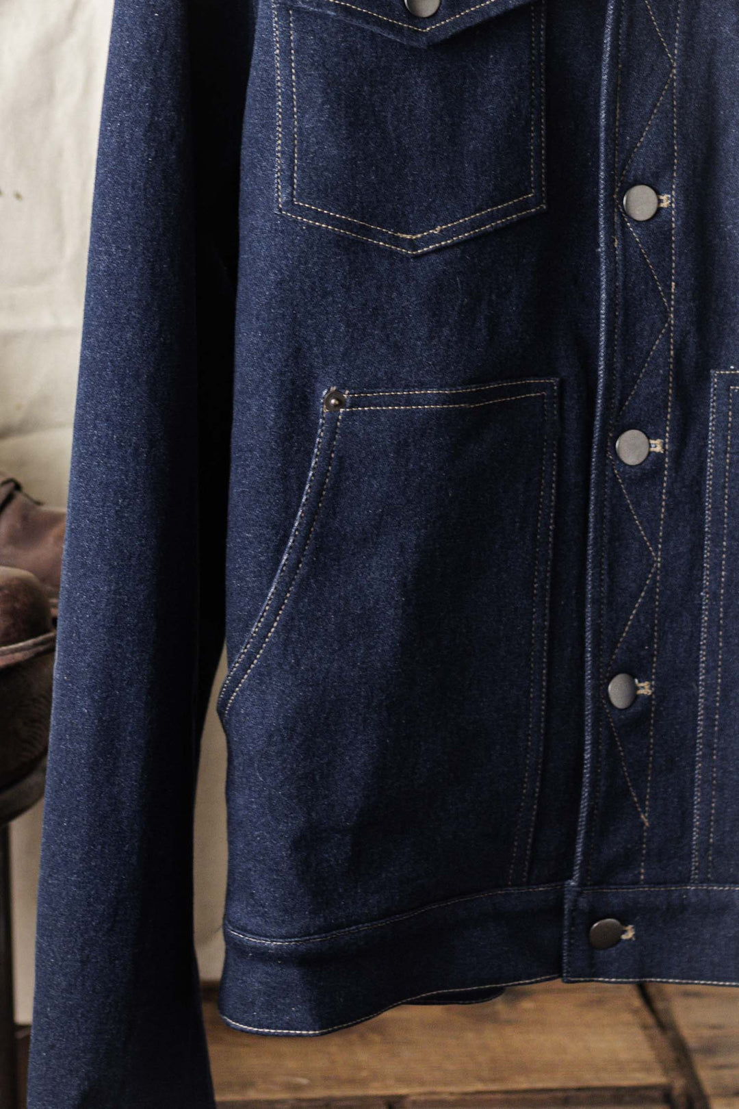 A close-up of the Bridge & Burn Burl Trucker Jacket in dark denim, featuring silver buttons, visible white stitching, chest and lower pockets, and part of the sleeve.