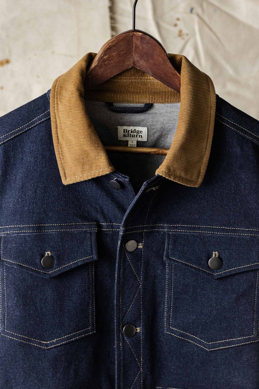 The Bridge & Burn Burl Trucker Jacket in Dark Denim hangs on a wooden hanger, showcasing a brown corduroy collar, two buttoned chest pockets, and white contrast stitching.