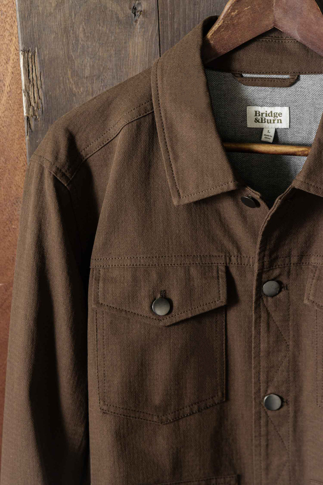 The Bridge & Burn Burl Trucker Jacket in Walnut hangs on a wooden hanger against a rustic wooden backdrop, featuring flap chest pockets and a visible inner collar tag with the brand name.