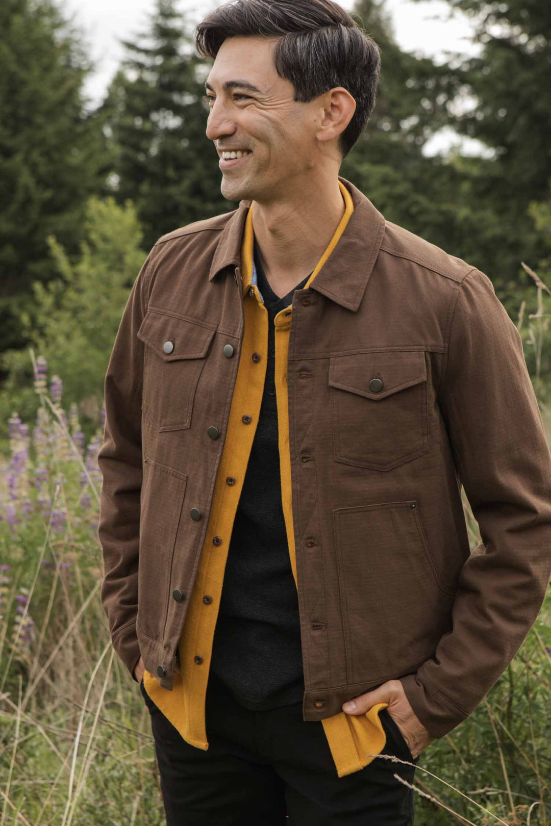 A man stands smiling outdoors among trees, tall grass, and flowers. He wears the Bridge & Burn Burl Trucker Jacket in Walnut over a yellow shirt and dark sweater.