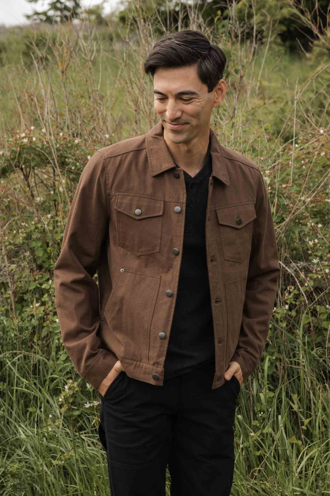 A man stands outdoors before tall grass, wearing the Bridge & Burn Burl Trucker Jacket in Walnut over a black shirt. With dark hair and hands in pockets, he looks down with a slight smile.