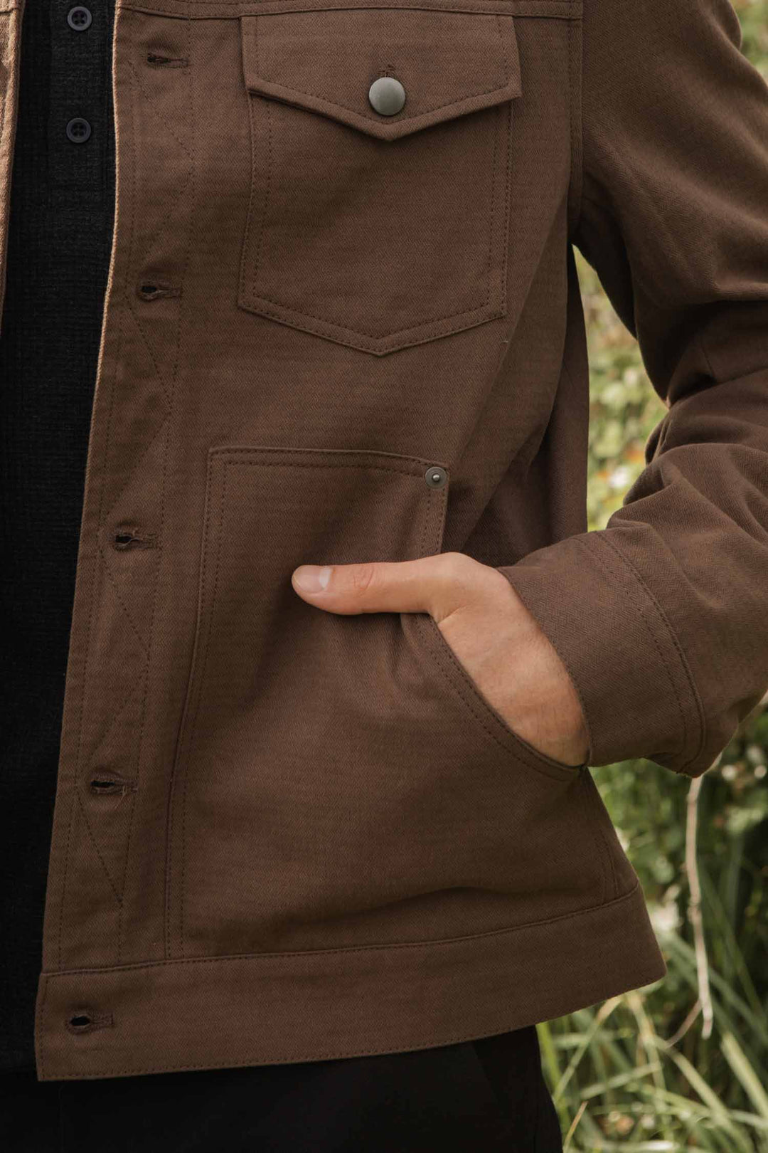 A person wearing the Bridge & Burn Burl Trucker Jacket in Walnut with flannel lining and one hand in the front pocket, standing outdoors with greenery in the background.